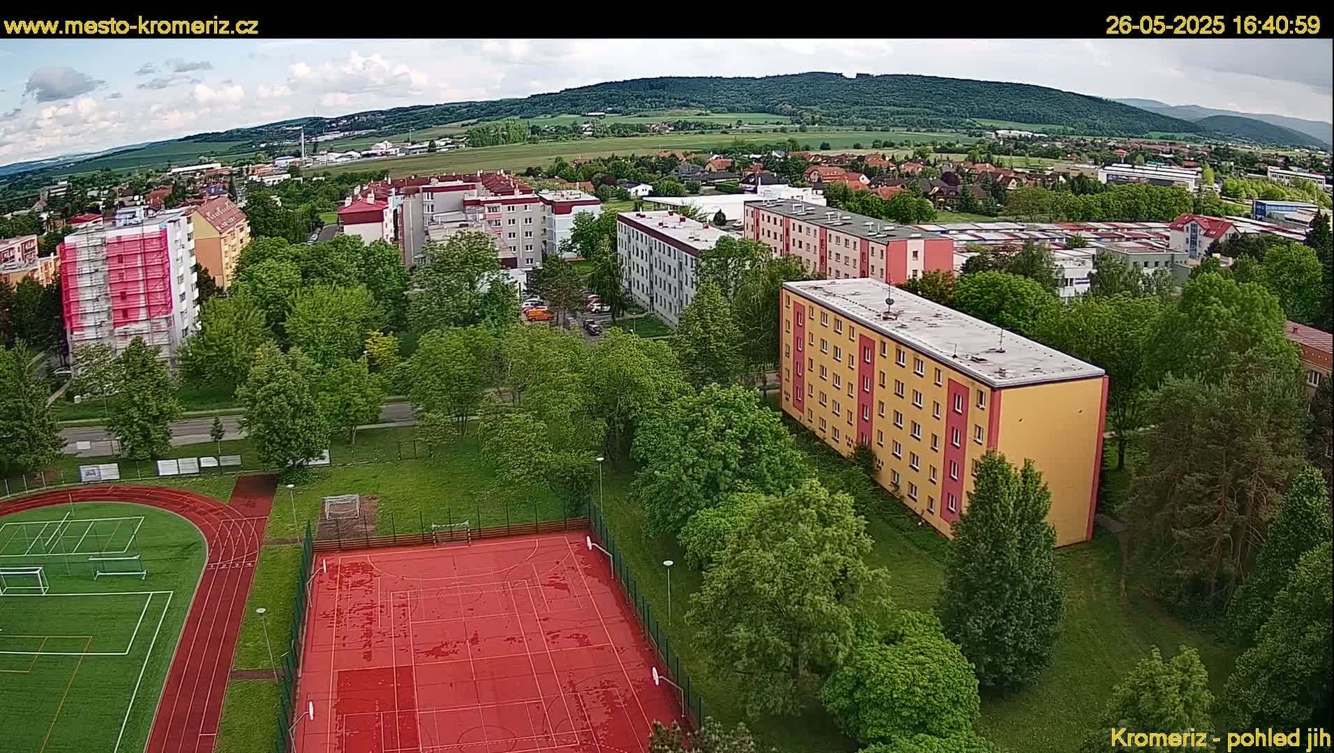 An aerial view shows a residential area with apartment buildings and a sports field, under partly cloudy skies.