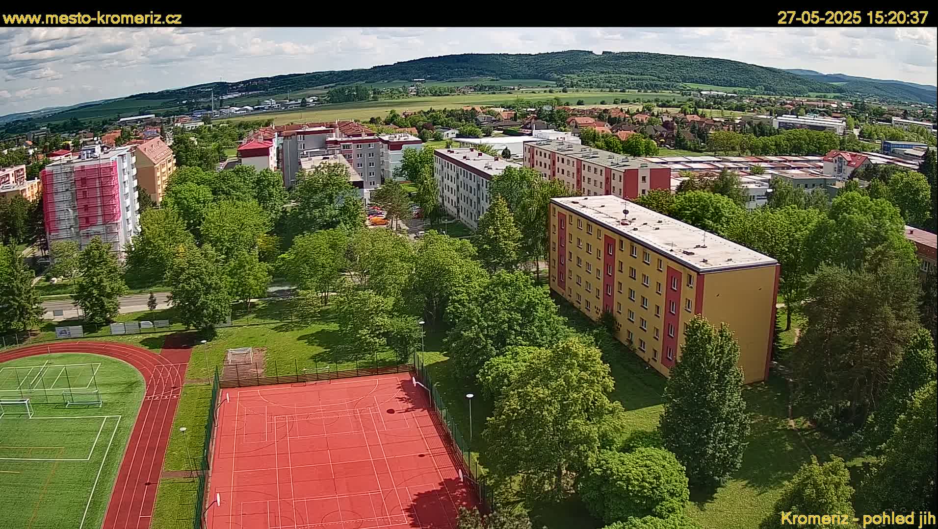 An aerial view shows a residential area with several multi-story apartment buildings surrounded by lush green trees and a red outdoor sports court next to a running track under a partly sunny sky.