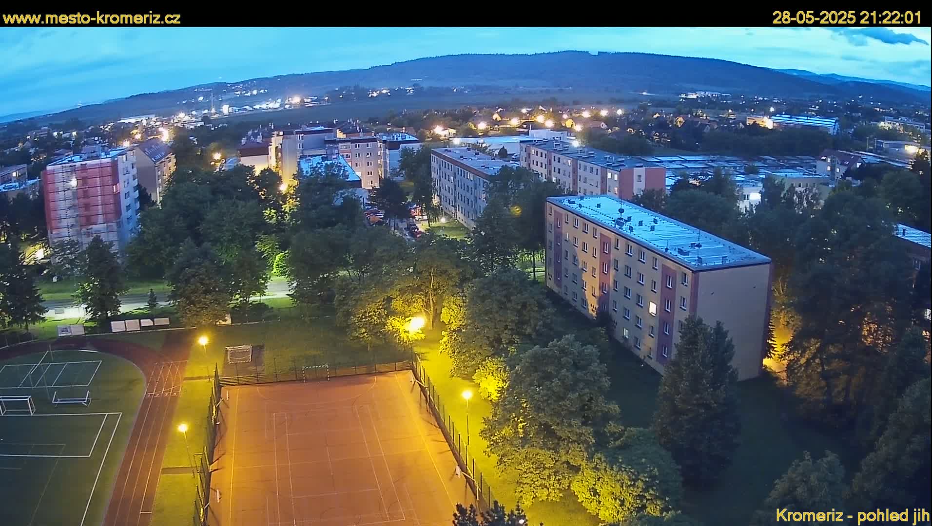An aerial nighttime view of an apartment complex and sports fields illuminated by streetlights under a twilight sky.