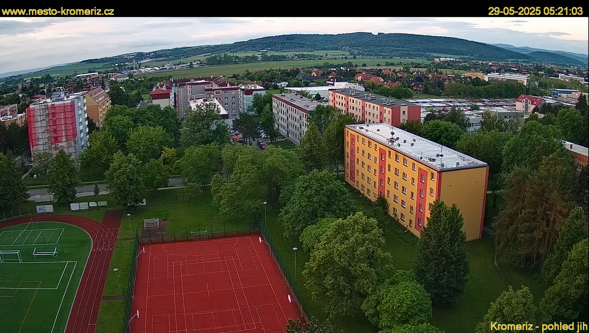 An aerial view shows a residential area with apartment buildings, green spaces, and a sports field under partly cloudy skies.