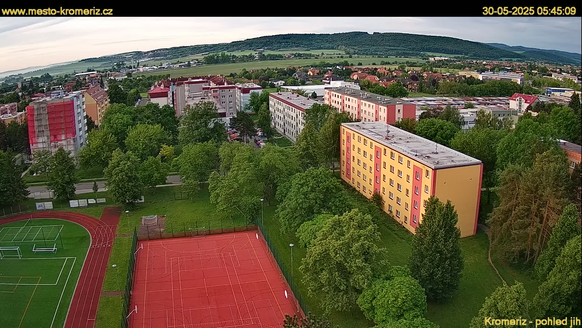 An aerial view shows a residential area with apartment buildings surrounded by lush green trees and a red sports court with a running track under a partly cloudy sky.