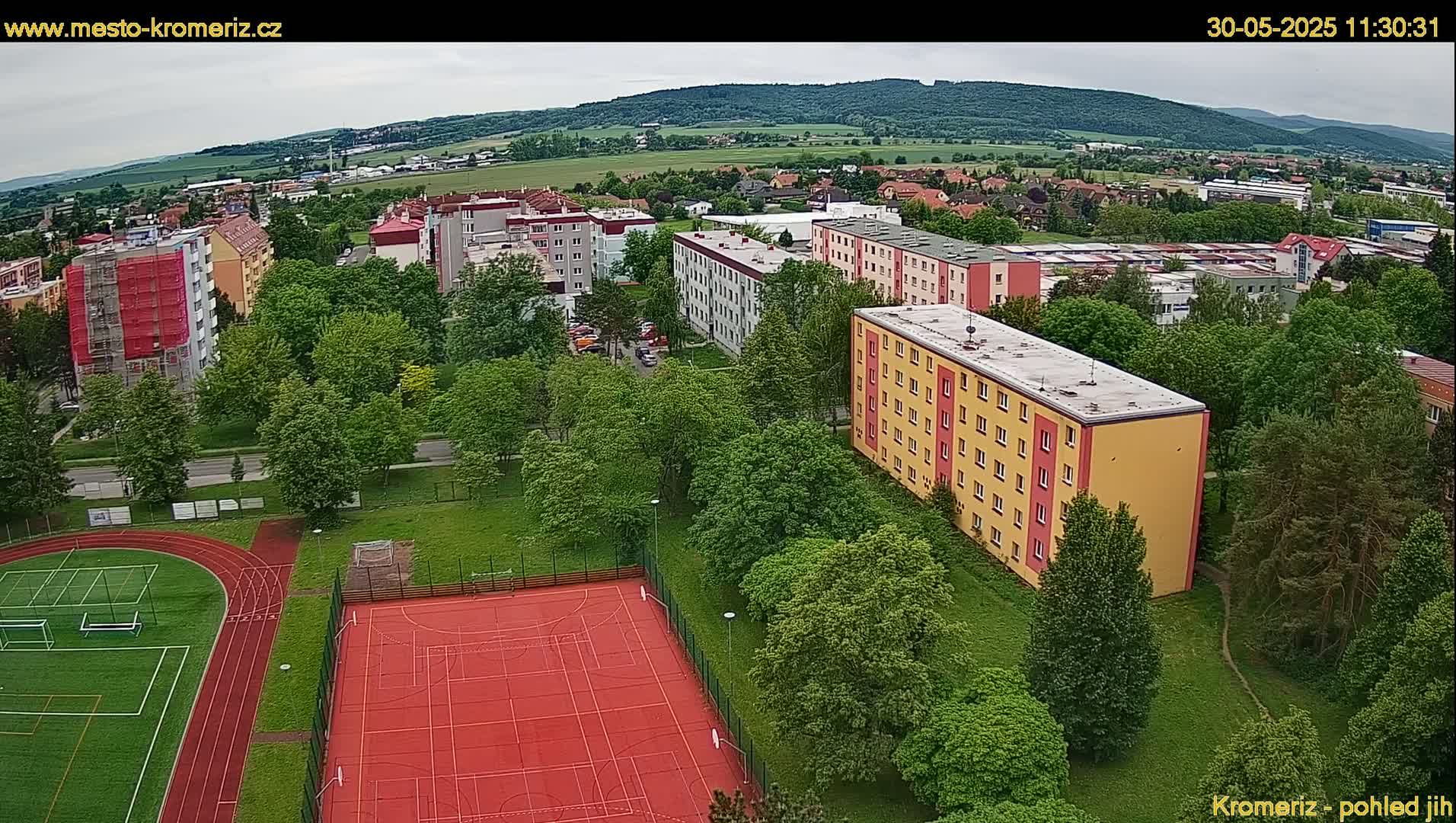 An aerial view shows a residential area with apartment buildings surrounded by lush green trees and a sports field with a running track and a basketball court under an overcast sky.