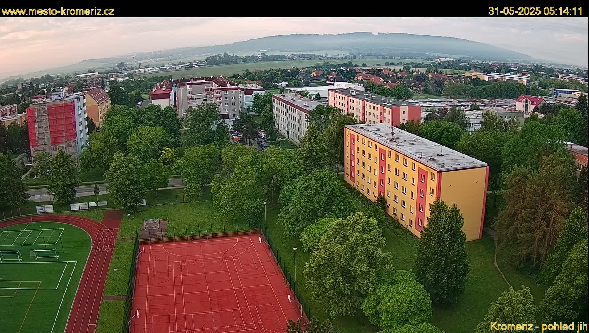 An aerial view shows a residential area with apartment buildings, a grassy area with lush green trees, and a red sports court with a running track under a partly cloudy sky.