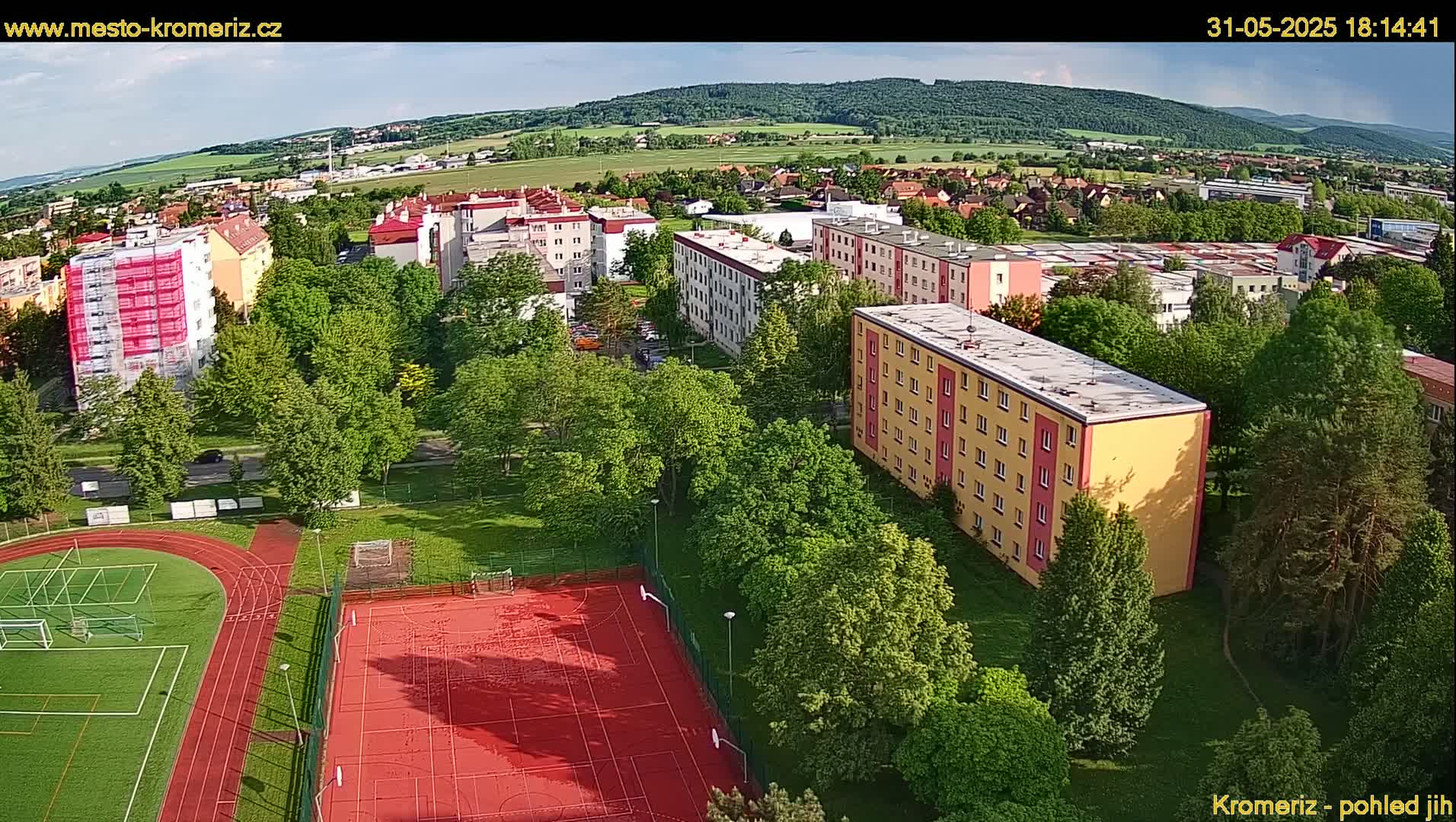 An aerial view shows a residential area with multi-story buildings, green trees, and athletic fields, under a partly sunny sky.