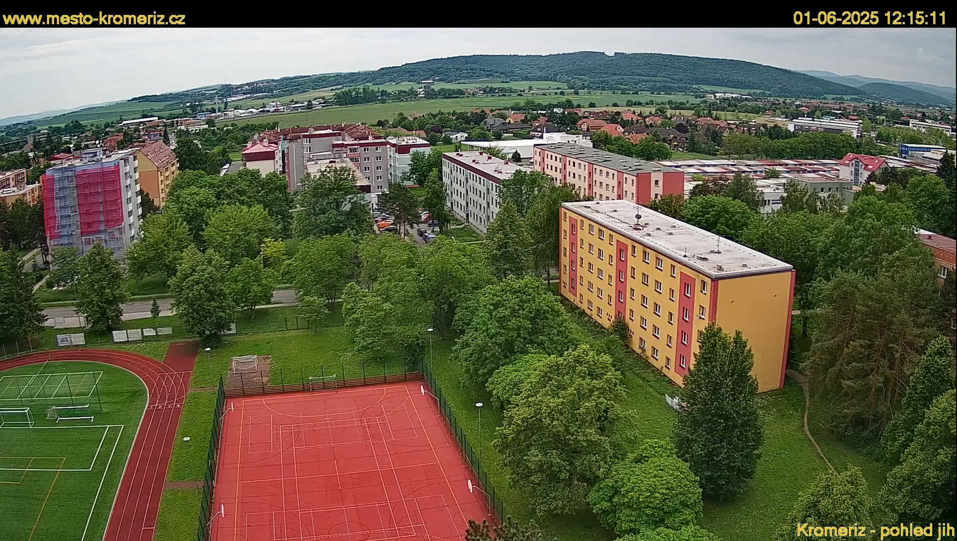 An aerial view shows an apartment complex, a running track, and a multi-use sports court surrounded by lush green trees under a partly cloudy sky.