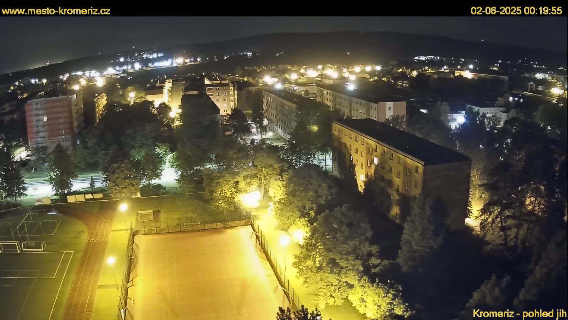 A nighttime aerial view shows a residential area with illuminated buildings, trees, and a sports field, under a clear, dark sky.