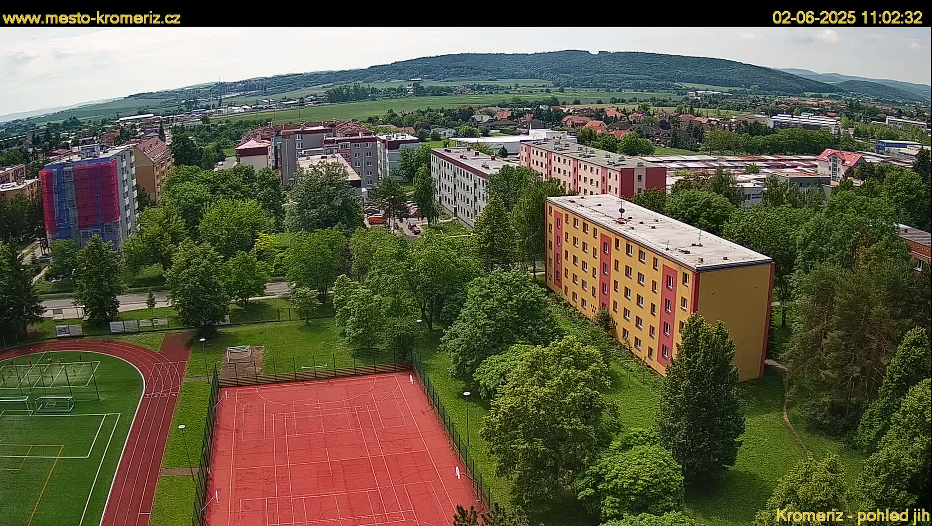 An aerial view shows a residential area with apartment buildings surrounded by lush green trees and a sports complex with a running track and a red basketball court, under a mostly sunny sky.
