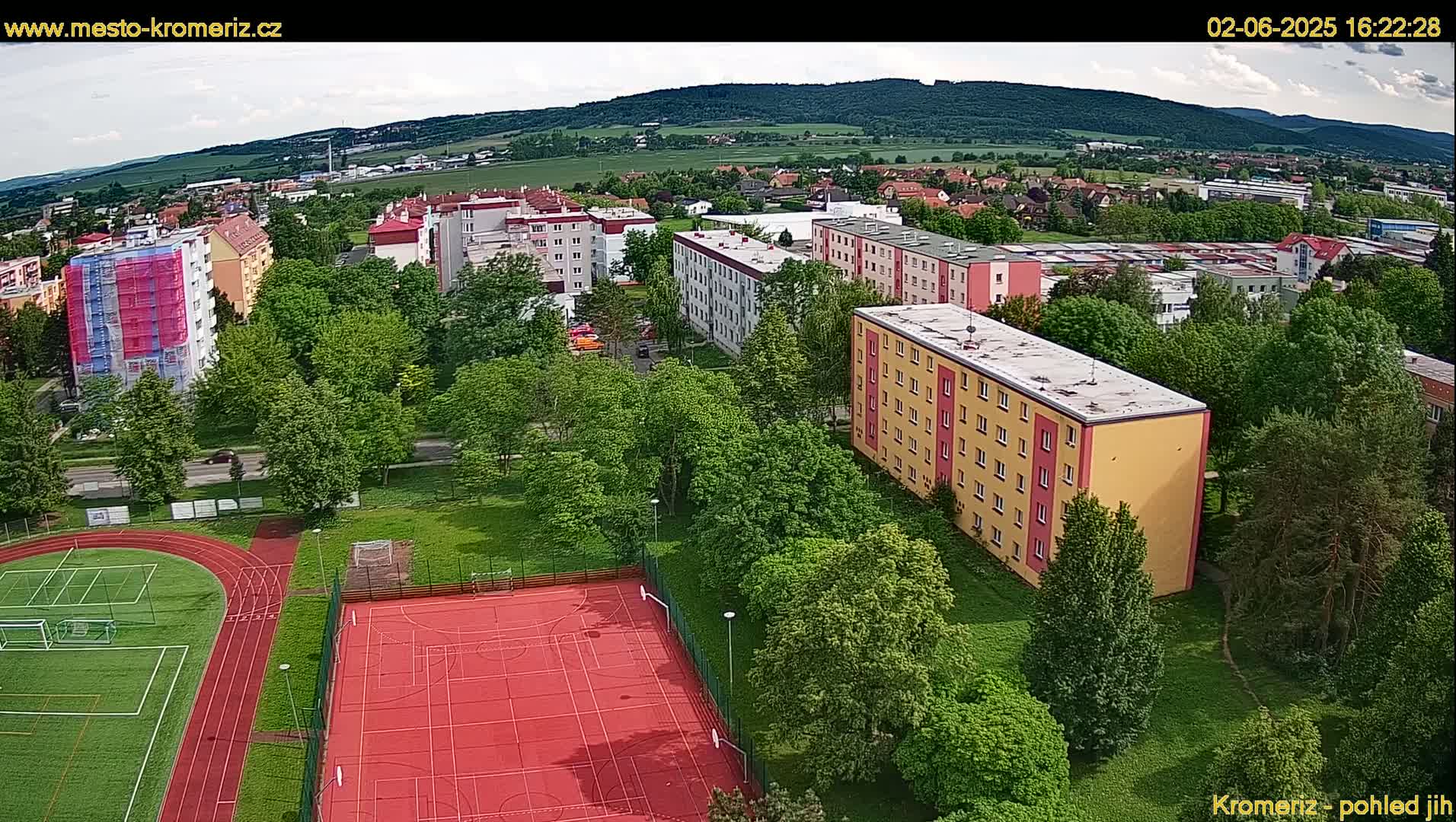 An aerial view shows a residential area with multi-story buildings, green trees, a running track, and a red multi-purpose sports court under partly cloudy skies.