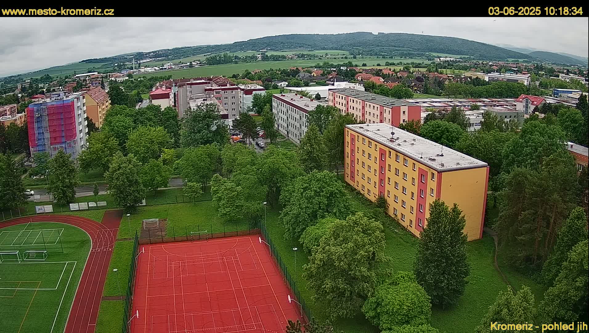 An aerial view shows a residential area with apartment buildings surrounded by lush green trees and a red sports court with a running track, under an overcast sky.