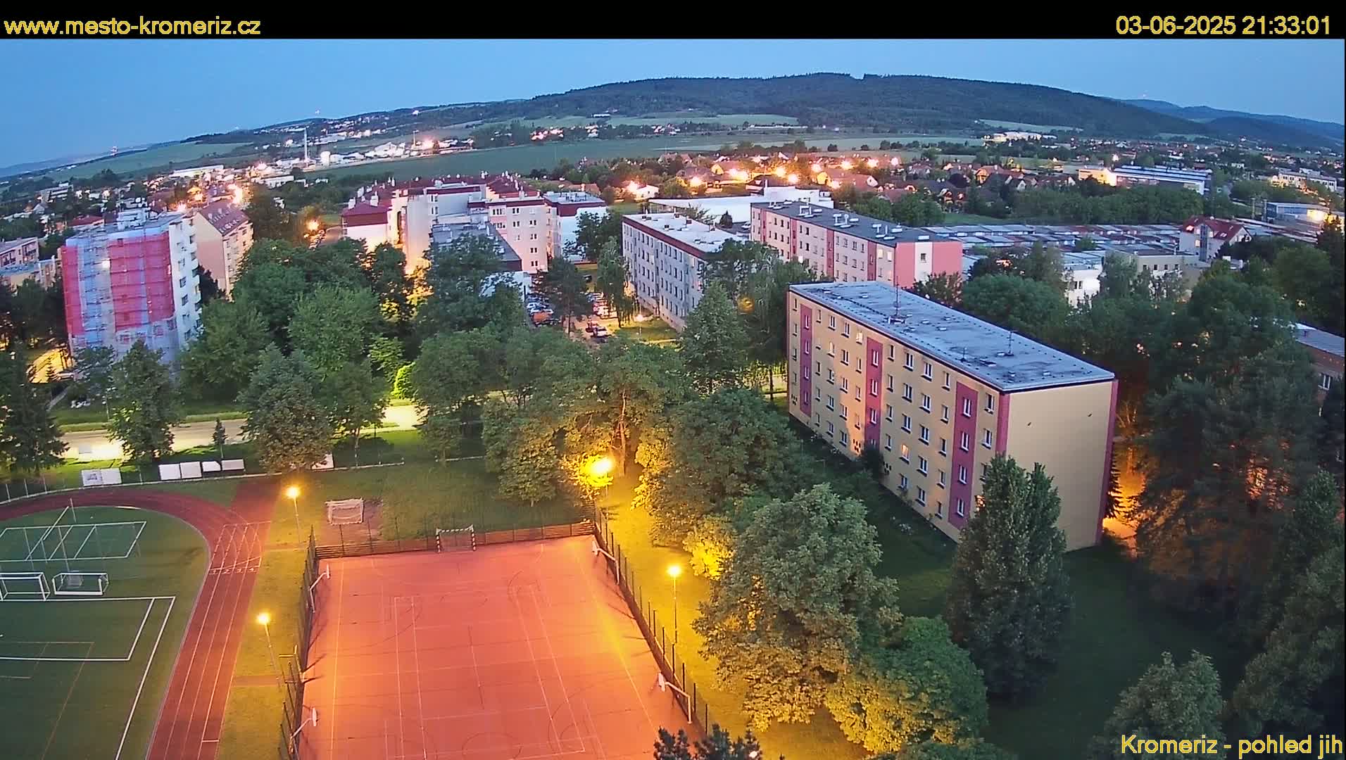 An aerial view at dusk shows a residential area with apartment buildings, a sports field with a running track, and trees, all under a clear sky.