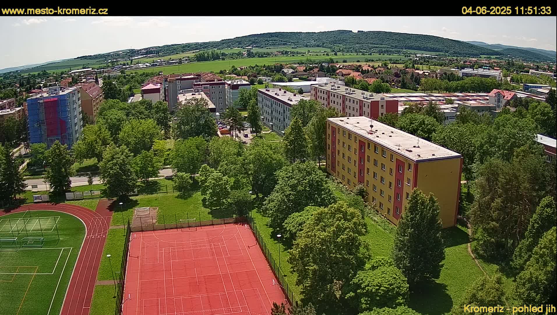 An aerial view shows a residential area with apartment buildings surrounded by lush green trees and a sports field with a running track and a red basketball court, under a sunny sky.