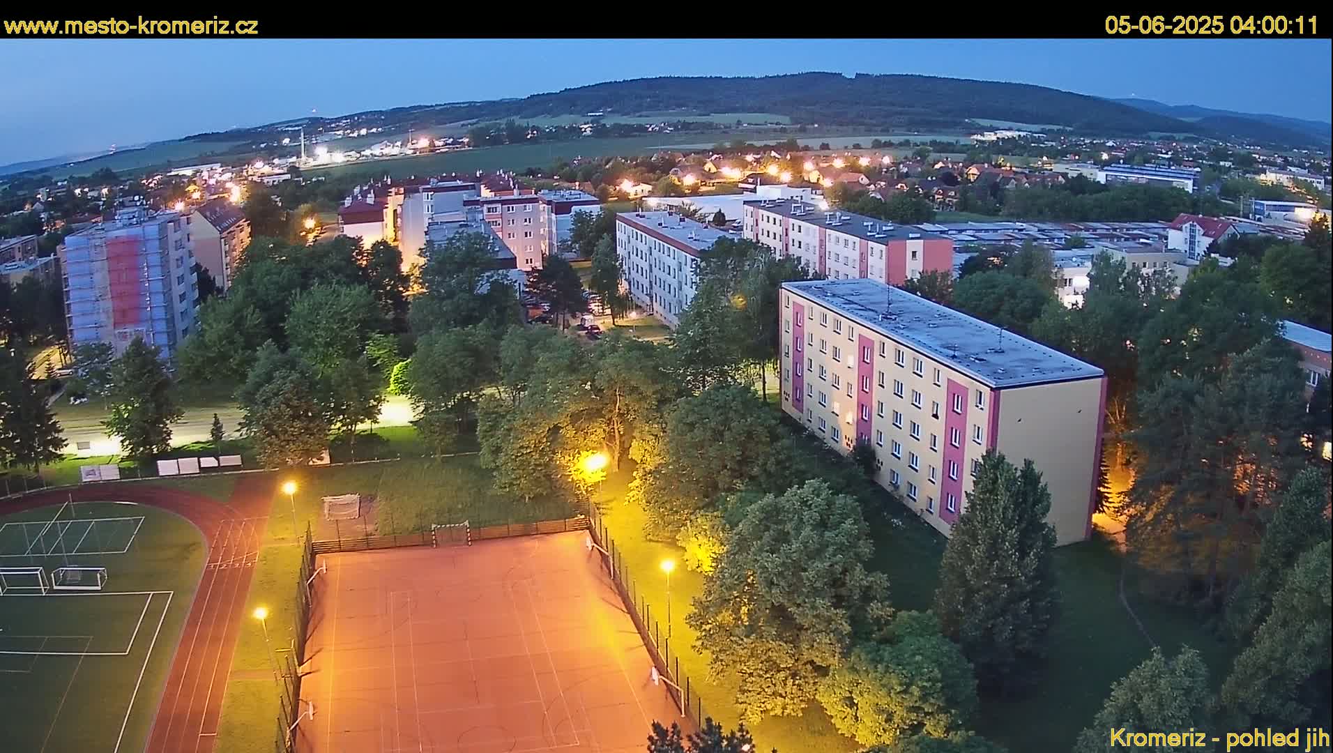 An aerial view at twilight shows a residential area with apartment buildings, green spaces, a running track, and a sports court, all illuminated by artificial lighting.