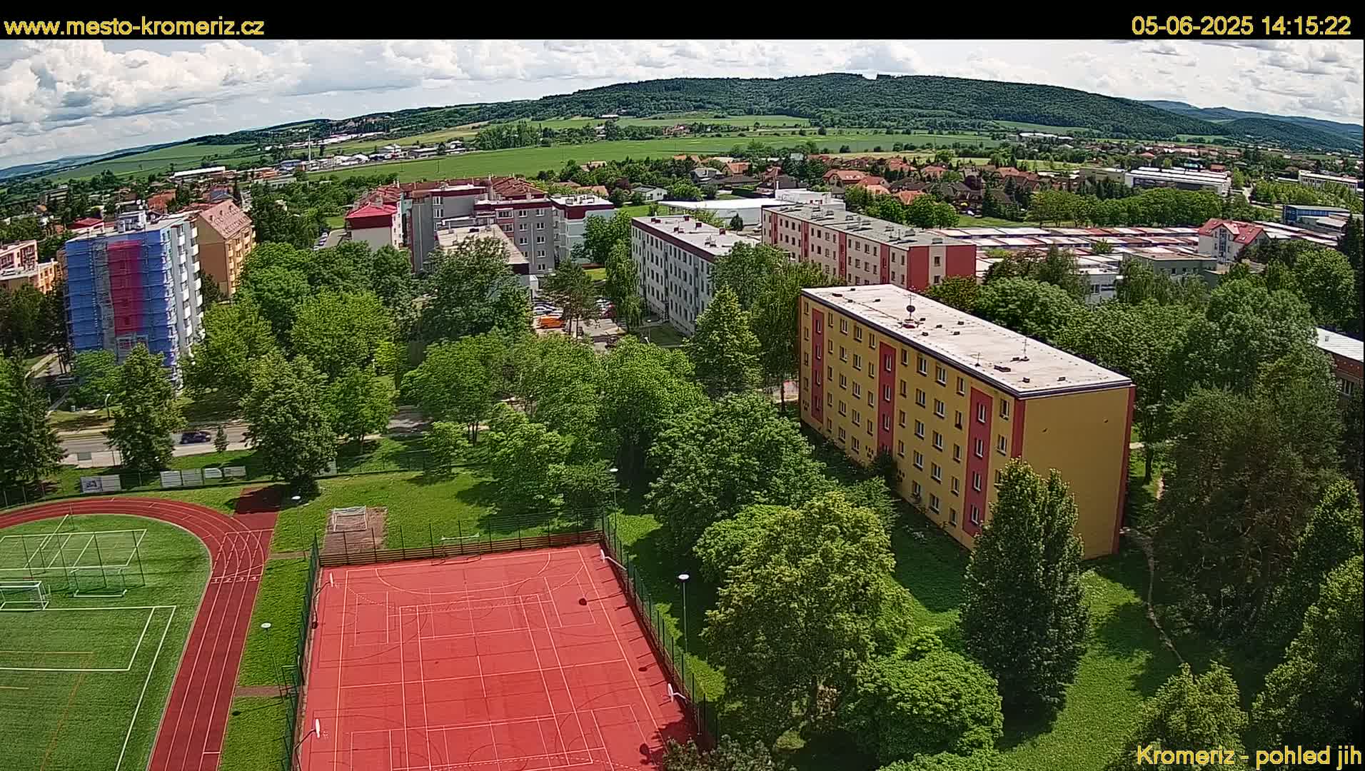An aerial view shows a residential area with apartment buildings, green spaces including a running track and multi-purpose sports court, under partly cloudy skies.