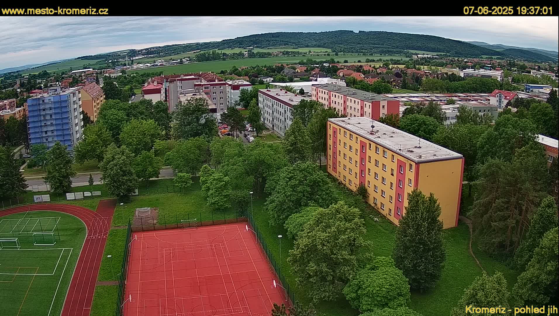 An aerial view shows apartment buildings, a running track, and a basketball court surrounded by lush green trees under a mostly cloudy sky.