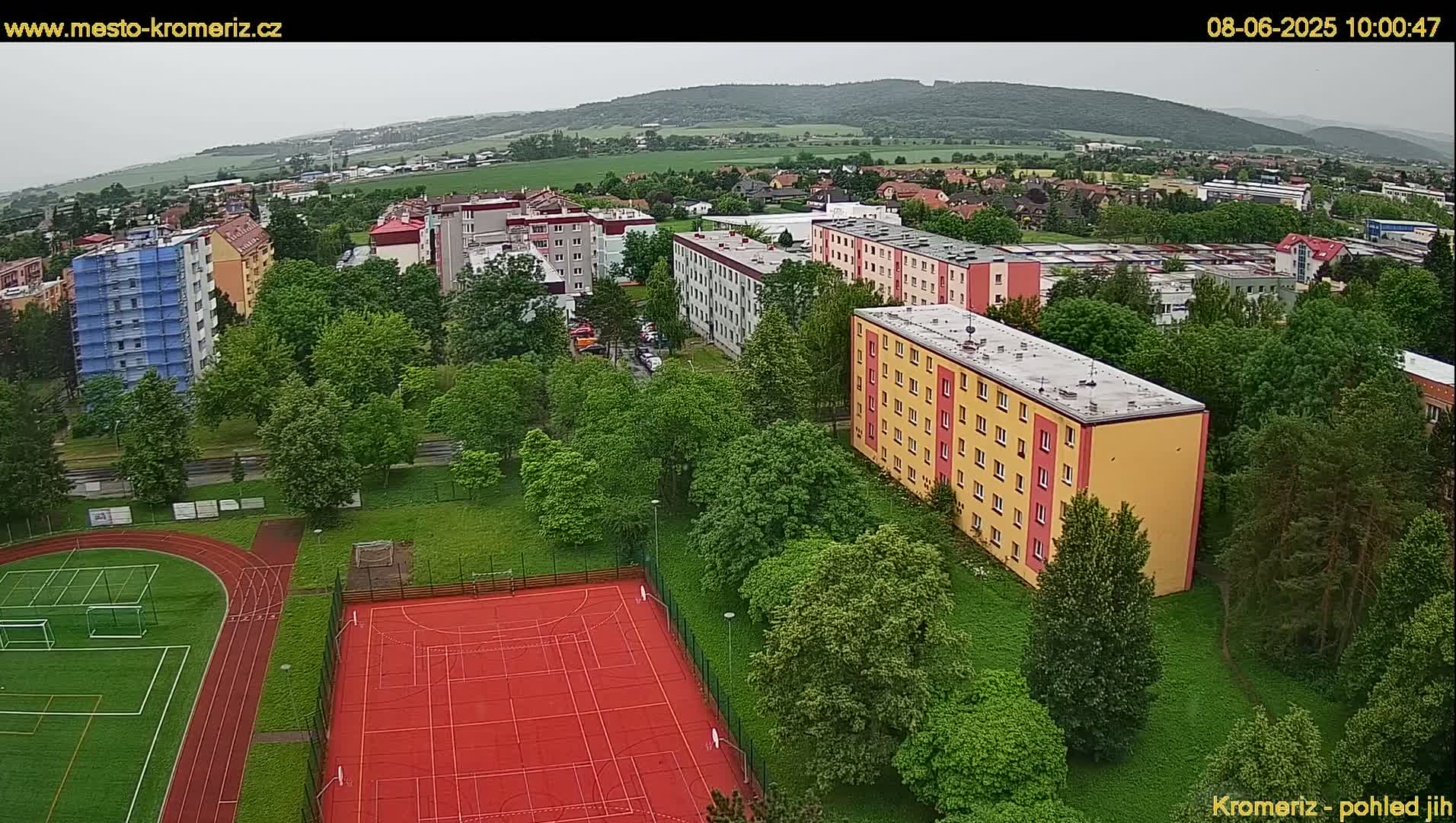 An aerial view shows an overcast day over a residential area with apartment buildings, green trees, and a red outdoor sports court with a running track.