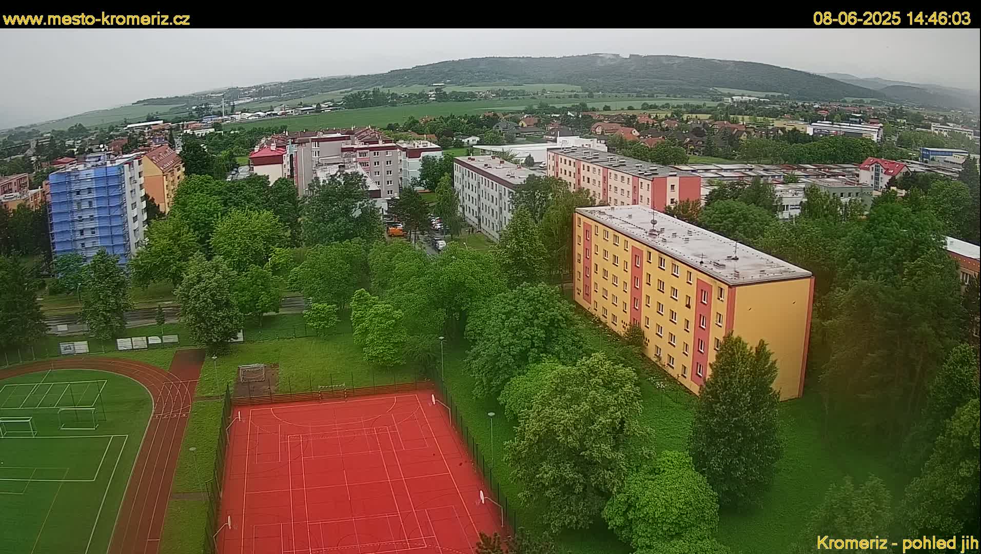 An aerial view shows an overcast day over a residential area with apartment buildings, green trees, a red sports court, and a running track.