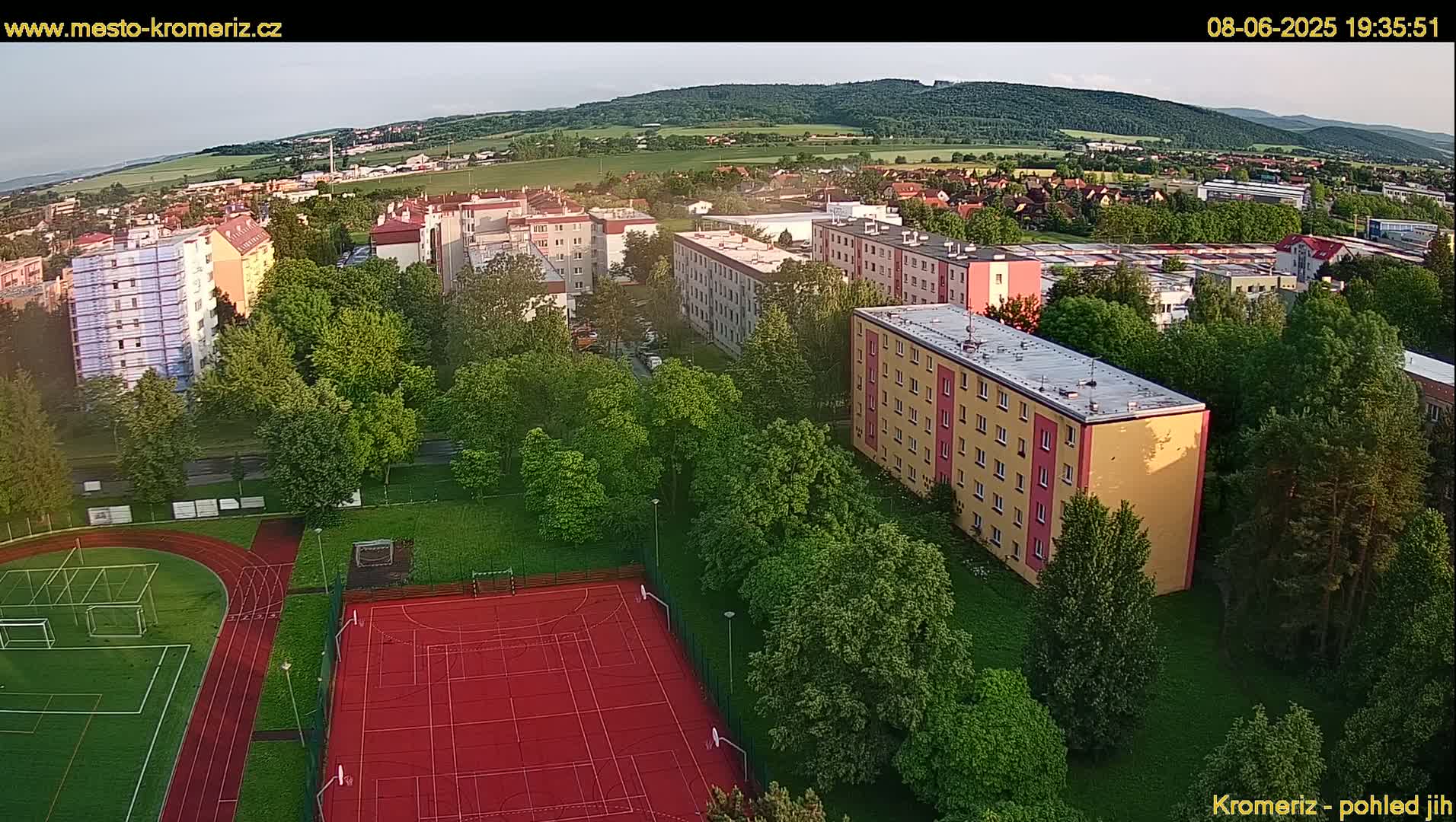 An aerial view of an apartment complex, a running track, and a sports court surrounded by lush green trees under a partly cloudy sky.