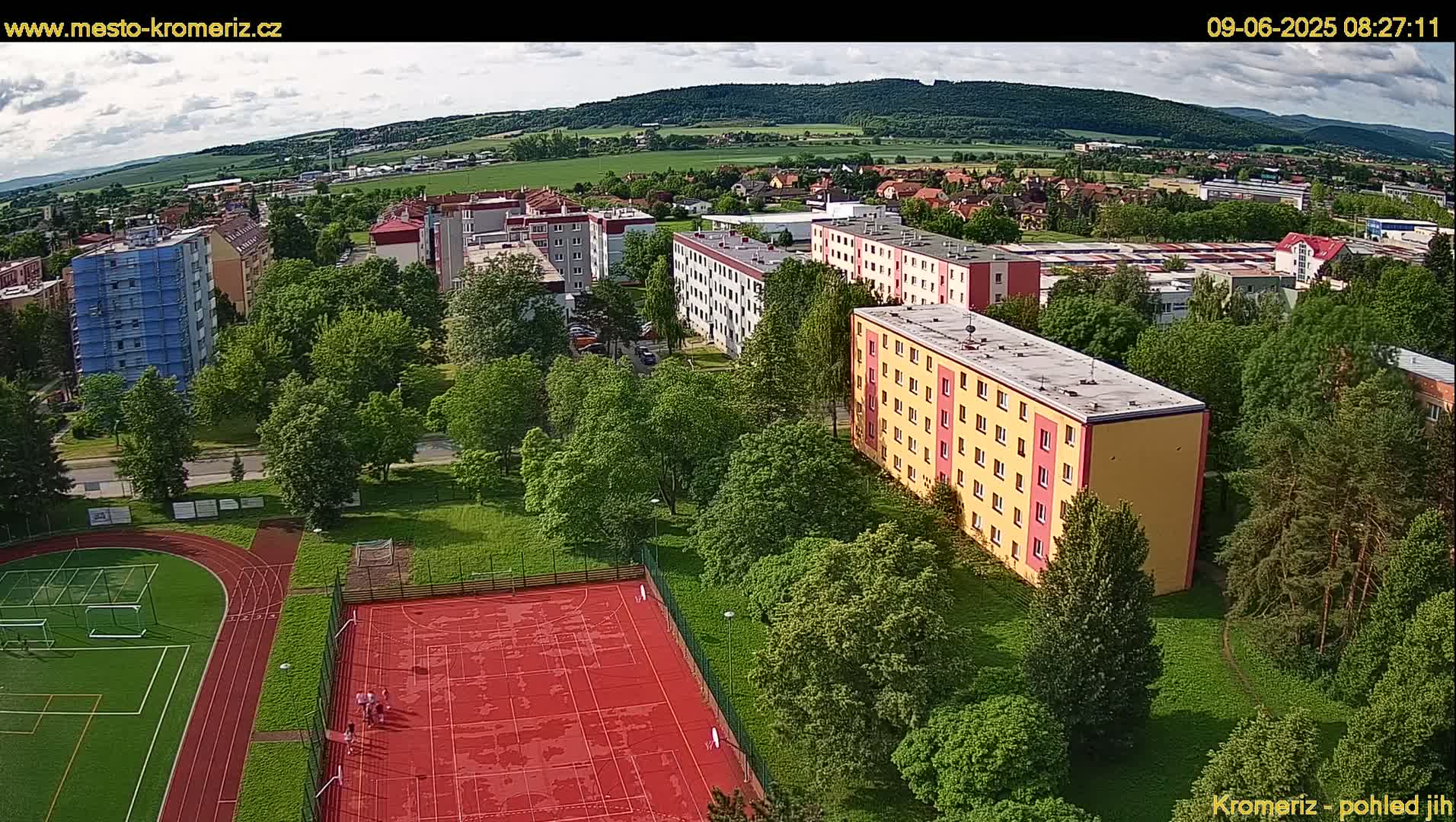 An aerial view shows several apartment buildings surrounded by lush green trees and a red sports court with a running track, under partly cloudy skies.
