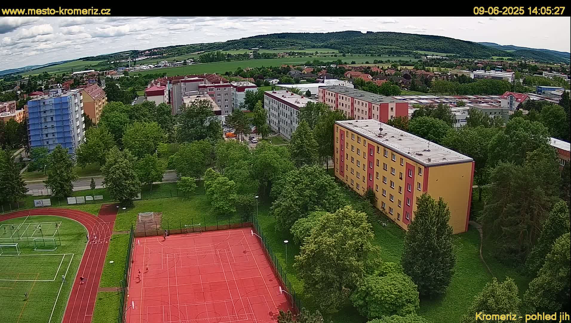 An aerial view shows a residential area with multi-story apartment buildings, a running track, and a basketball court, all surrounded by lush green trees under a partly cloudy sky.
