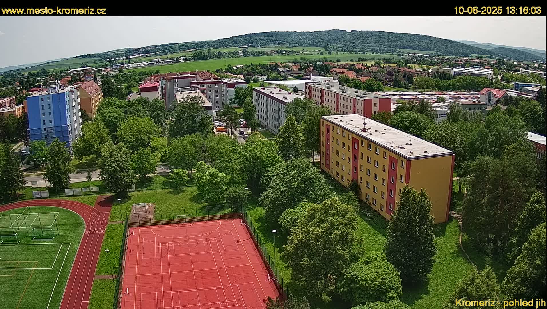 An aerial view shows apartment buildings, green spaces with trees, and a red sports court in a residential area on a sunny day.