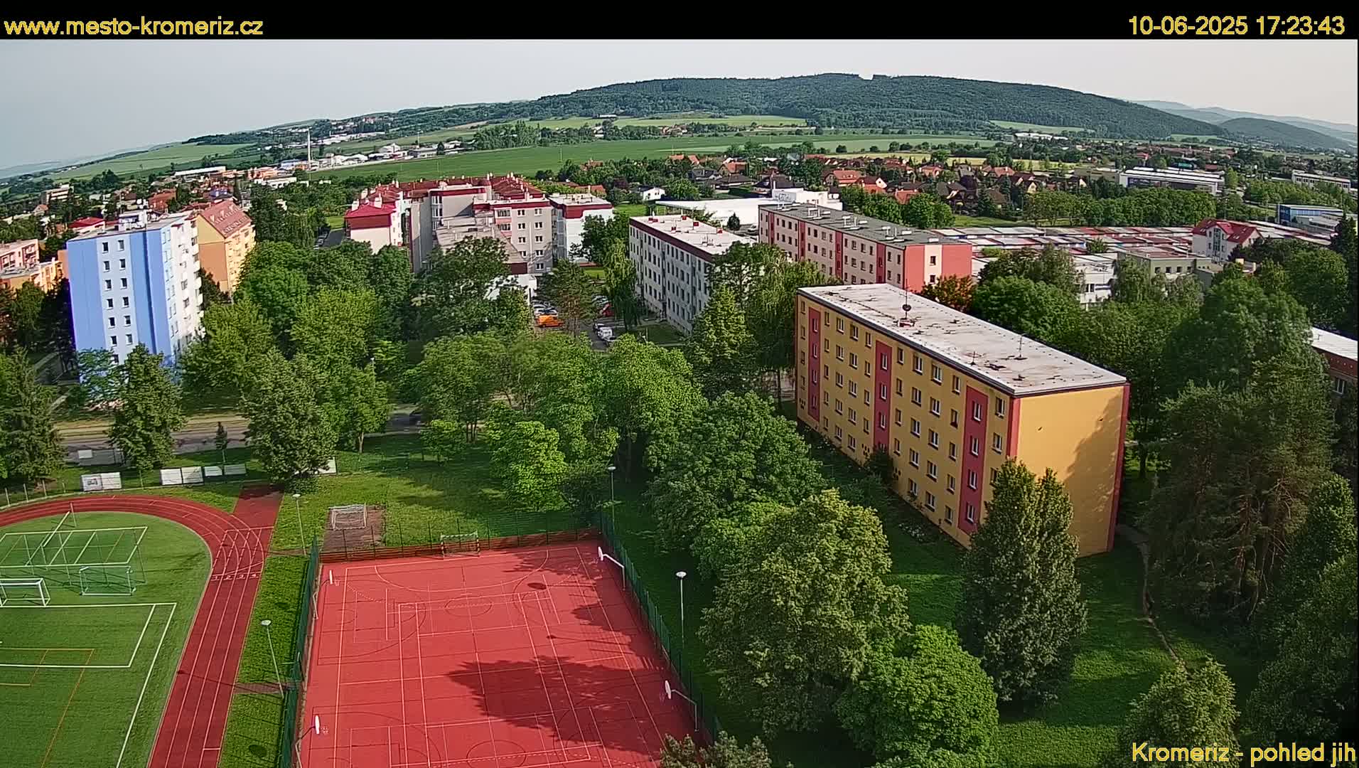 An aerial view of a residential area with apartment buildings, green spaces, and a red sports court, under a mostly clear sky.