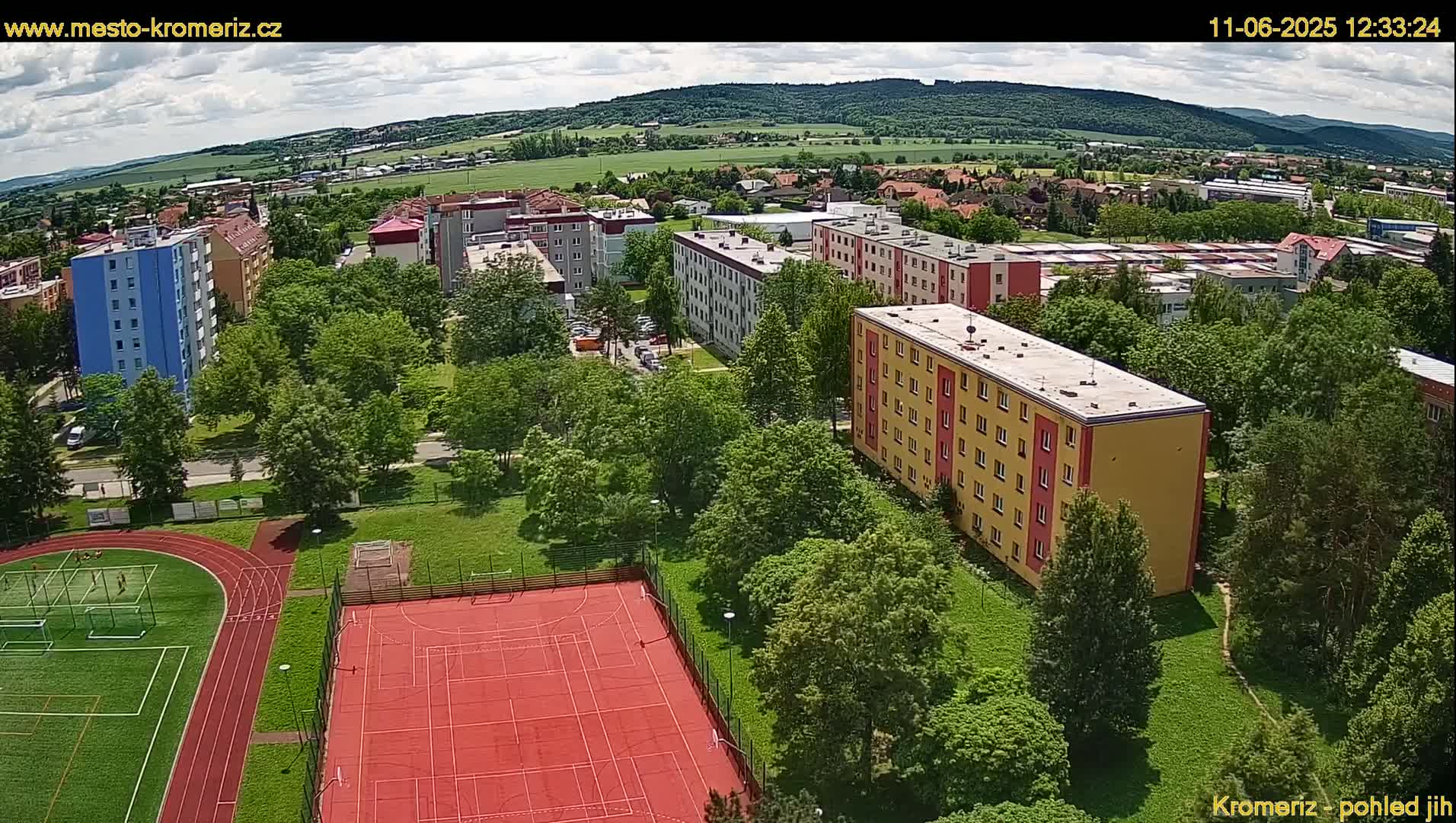 An aerial view shows a residential area with multi-story buildings surrounded by lush green trees and a red sports court, under a partly cloudy sky.