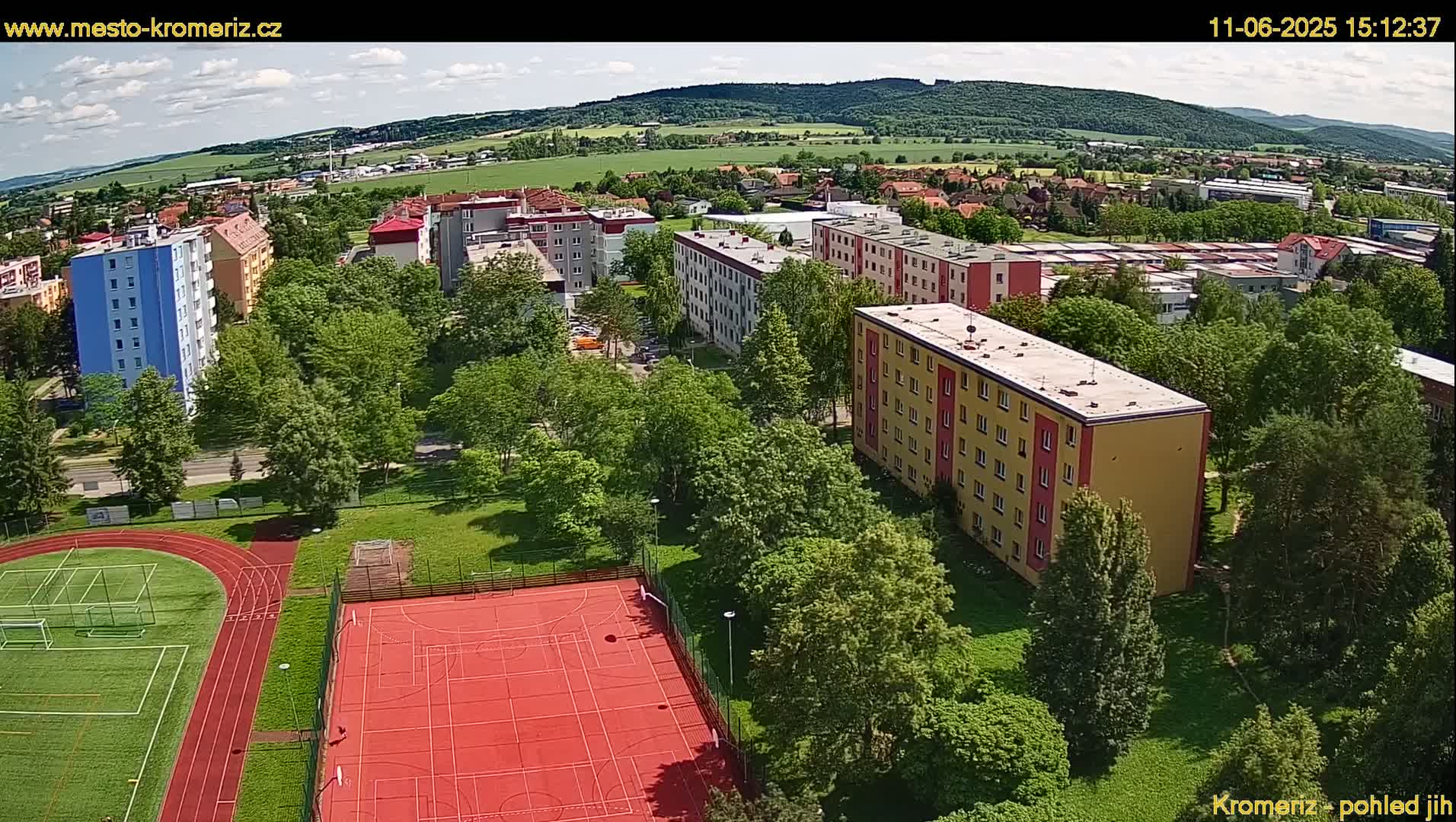 An aerial view of a residential area with apartment buildings, green spaces, a running track, and a multi-purpose sports court on a partly cloudy day.