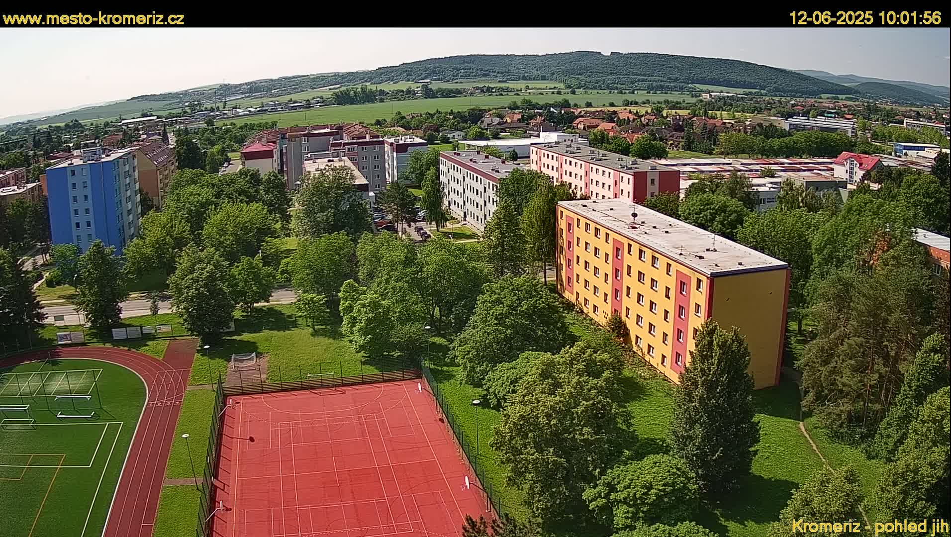 An aerial view shows a residential area with several apartment buildings, green trees, a running track, and a red sports court, all under a sunny sky.