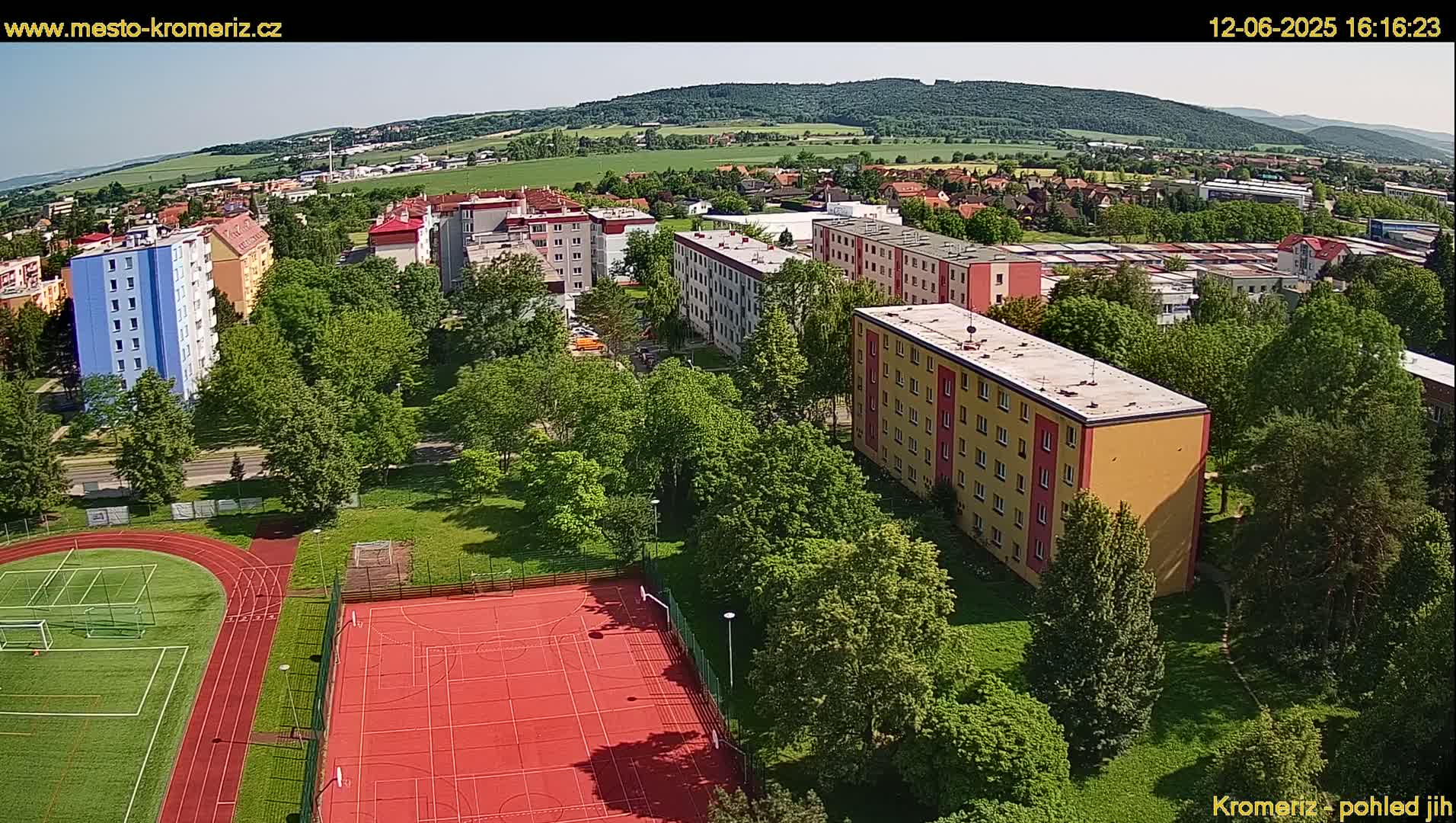 An aerial view shows a residential area with multi-story buildings, green spaces including a sports field with a running track and basketball court, under a sunny sky.