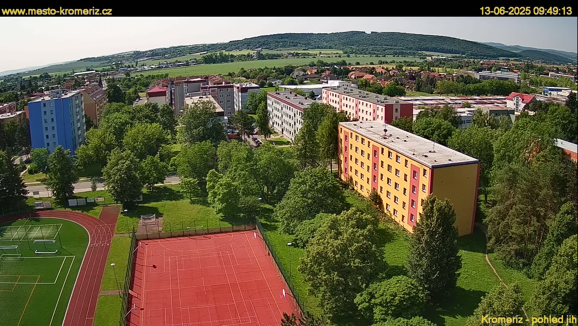 An aerial view shows several apartment buildings surrounded by lush green trees and a red outdoor sports court, on a sunny day.