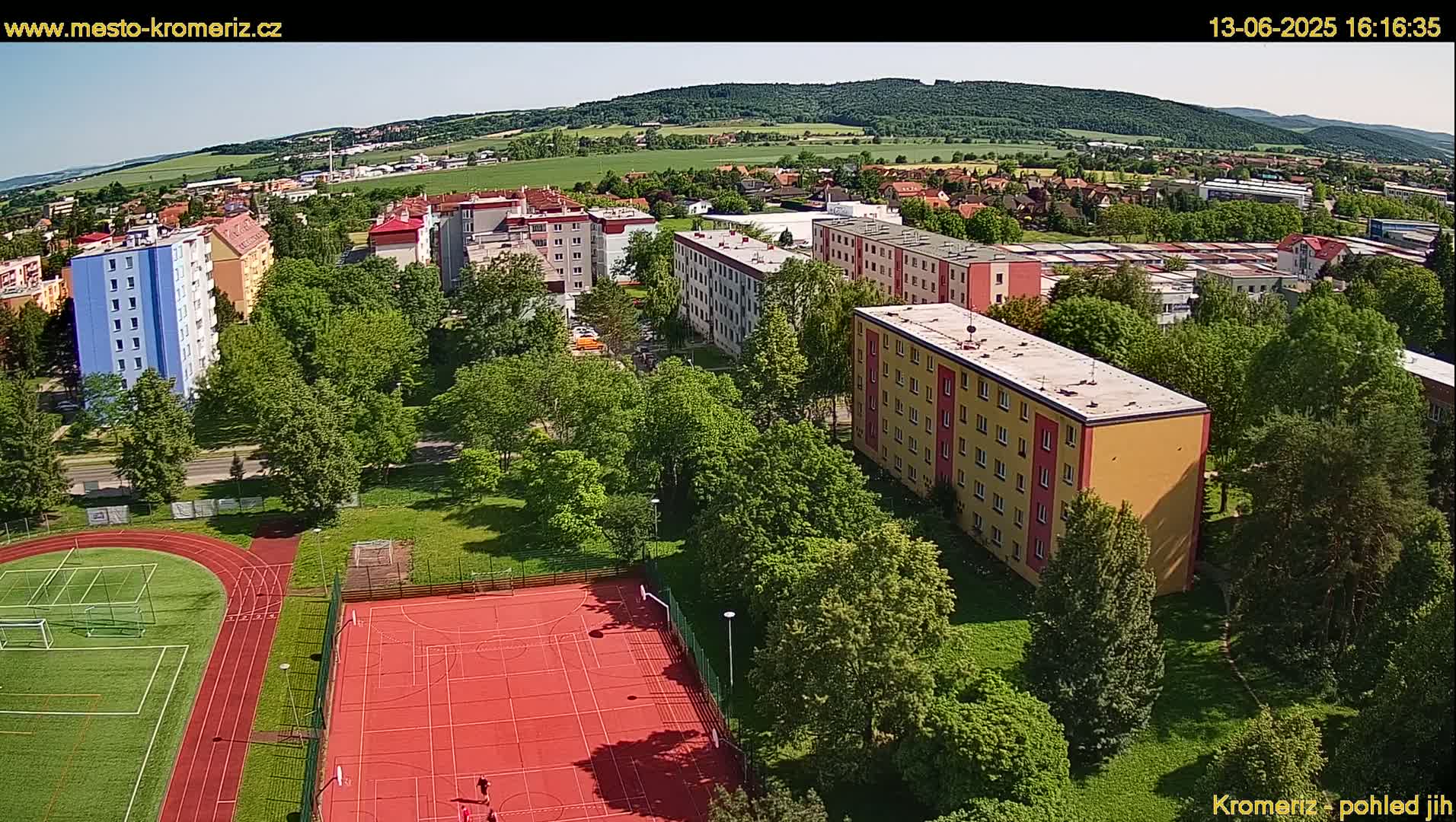 An aerial view shows a residential area with apartment buildings, green spaces, and a red sports court, on a sunny day.