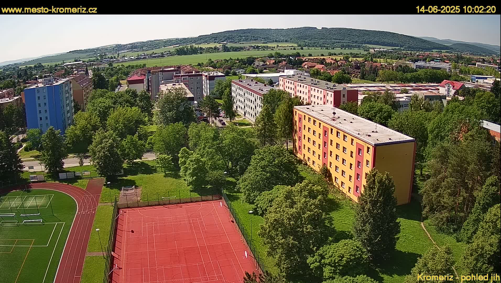 An aerial view shows a residential area with several apartment buildings surrounded by lush green trees and a multi-use sports field on a sunny day.
