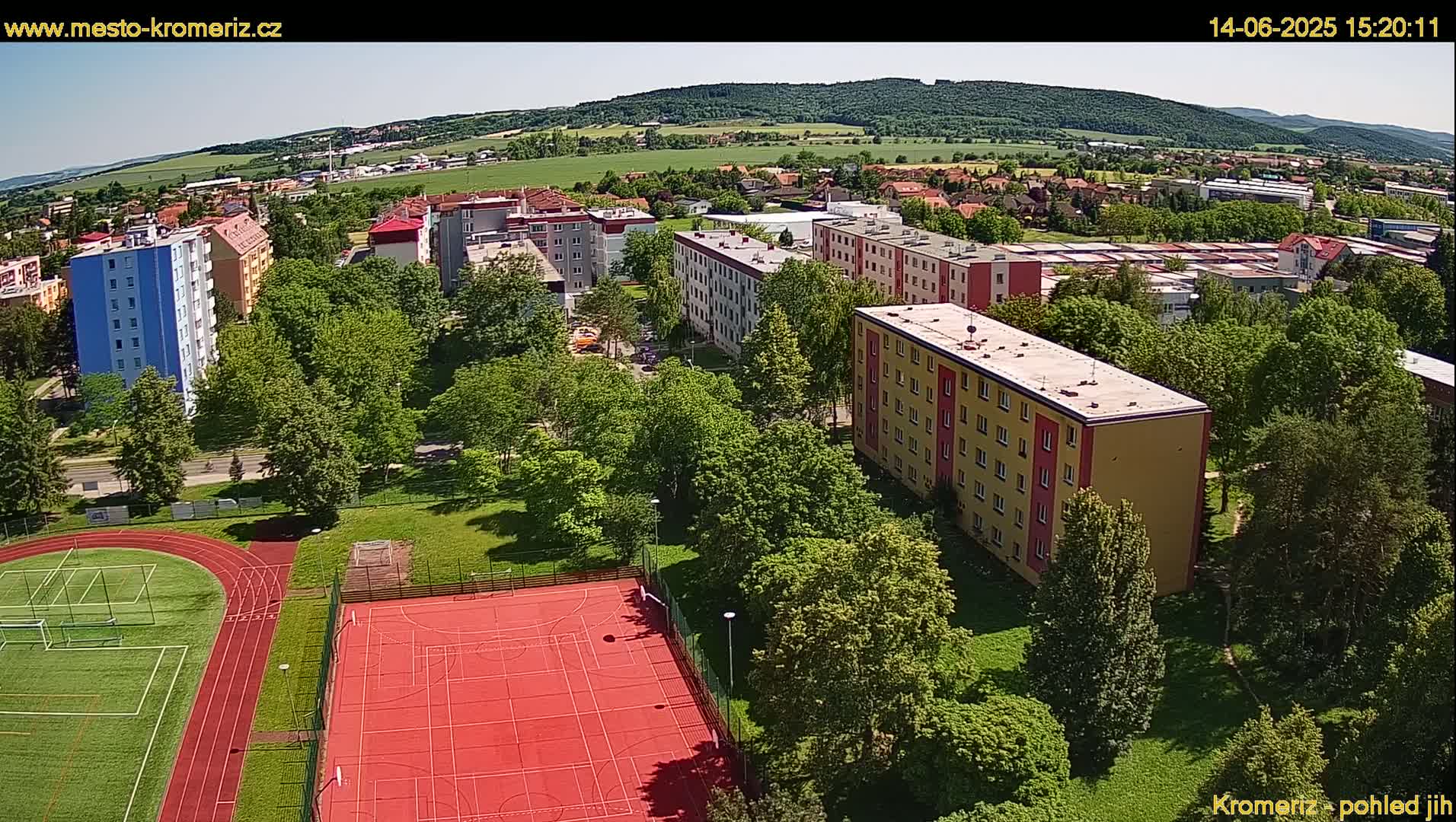 An aerial view shows a residential area with multi-story buildings and green spaces including a running track and a multi-purpose sports court, under sunny skies.
