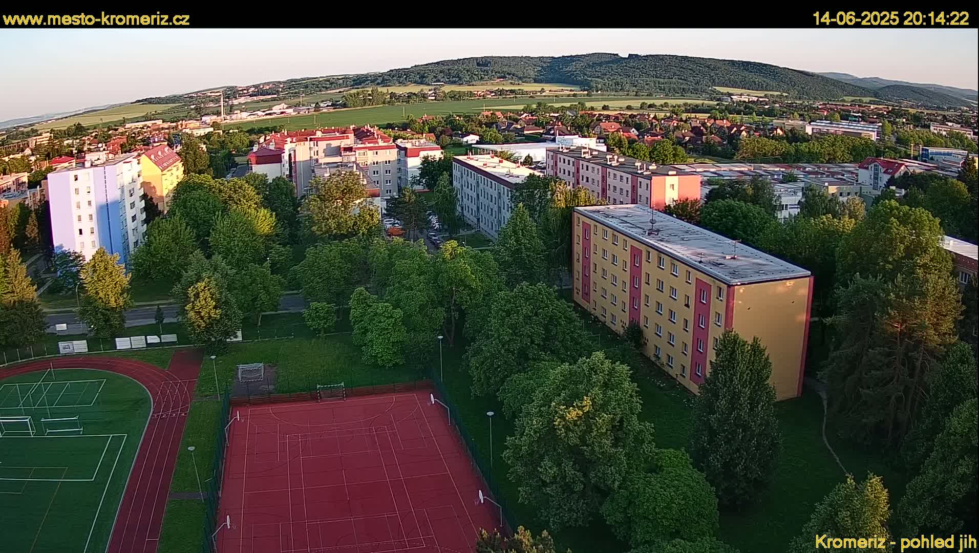 An aerial view shows a residential area with multi-story buildings and green spaces including a running track and a multi-purpose sports court, under sunny skies.