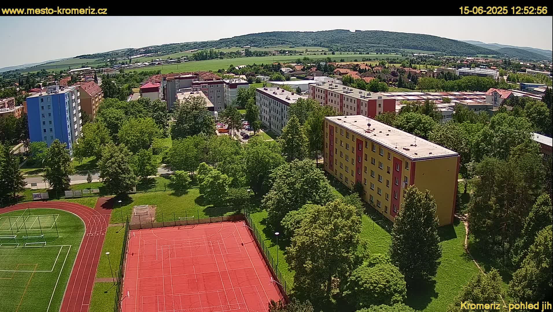 An aerial view of a residential area with several multi-story buildings surrounded by lush green trees and a red sports court, under a sunny sky.