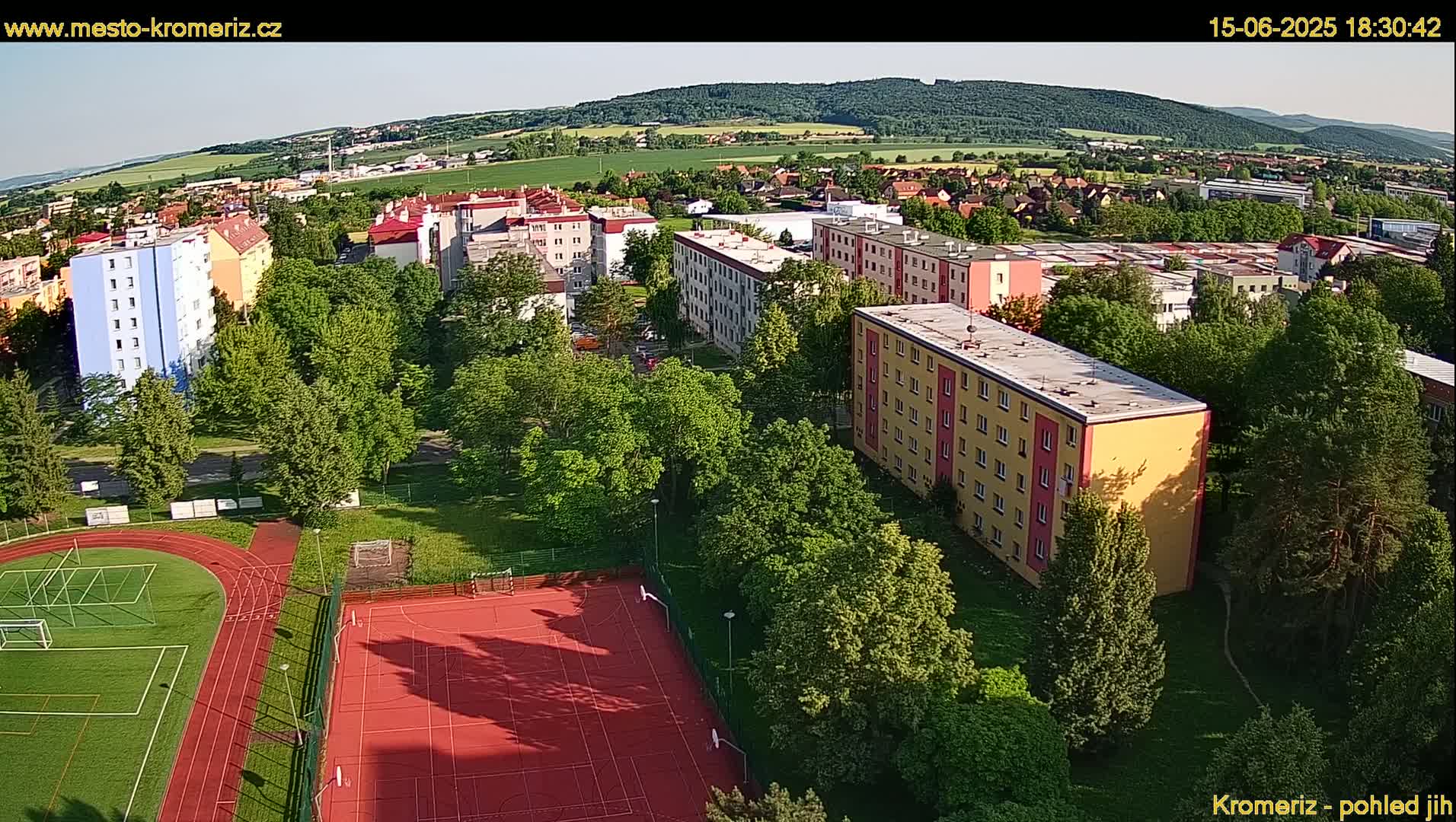 An aerial view shows a residential area with multi-story buildings surrounded by lush green trees and a sports field, under a clear daytime sky.