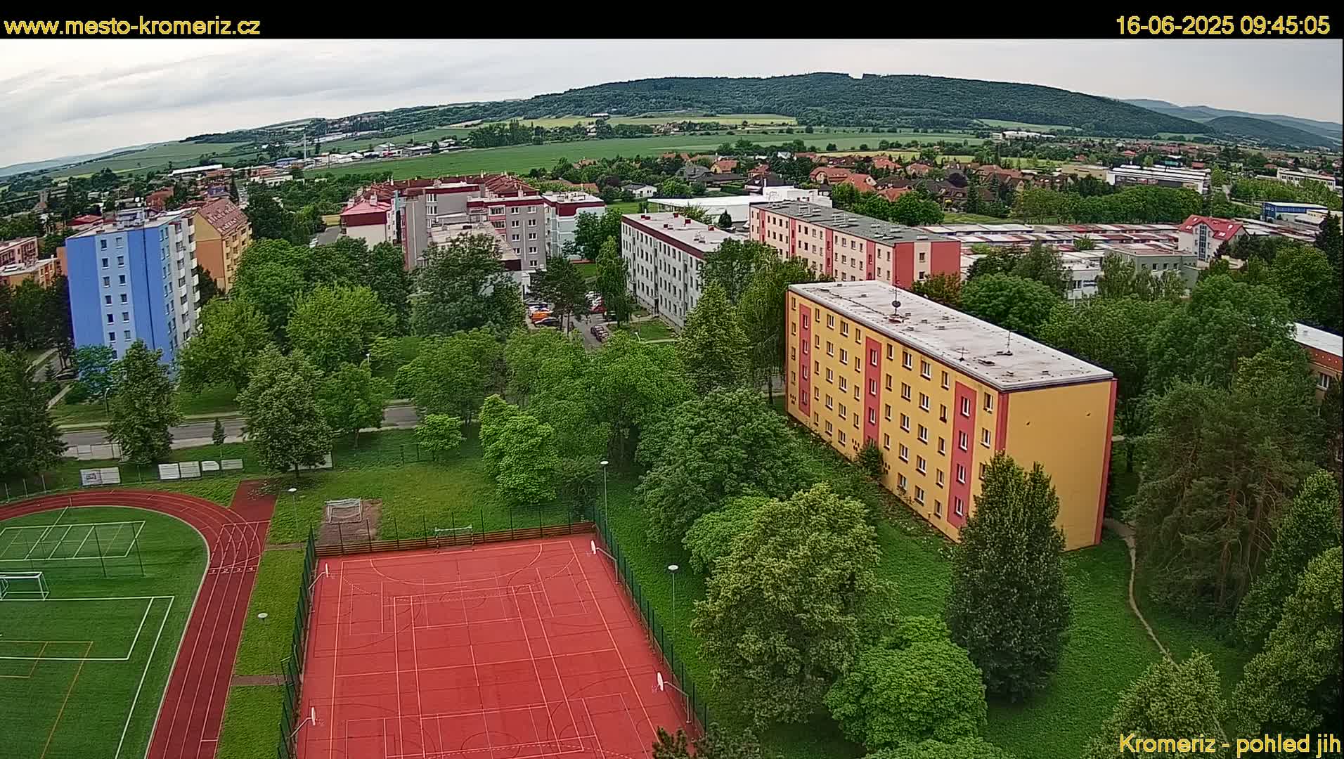 An aerial view shows a residential area with apartment buildings, green spaces, a running track, and a basketball court under an overcast sky.