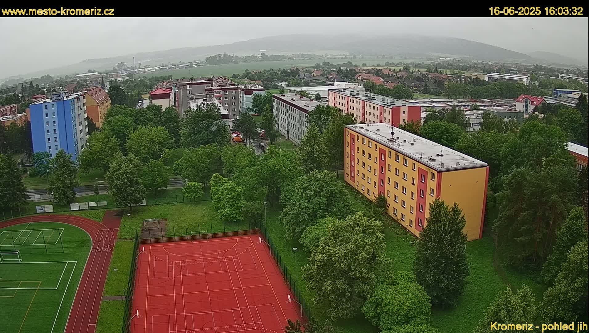 An aerial view shows a residential area with multi-story buildings, green spaces, and athletic fields, under an overcast sky.