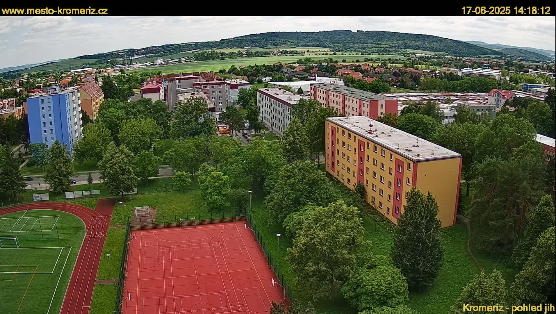 An aerial view shows several multi-story apartment buildings surrounded by green trees and a sports field with a running track under partly cloudy skies.