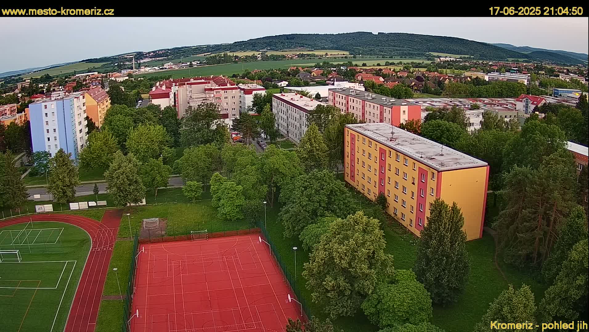 An aerial view shows multi-story apartment buildings surrounded by lush green trees and a red sports court with a running track, under a partly cloudy evening sky.