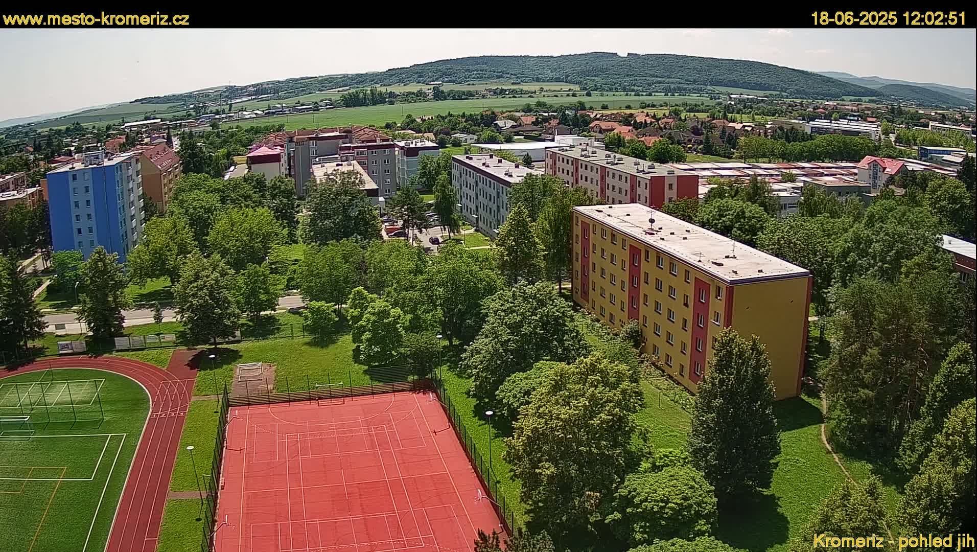 An aerial view shows a residential area with apartment buildings surrounded by green trees and a sports field with a running track and tennis court on a sunny day.