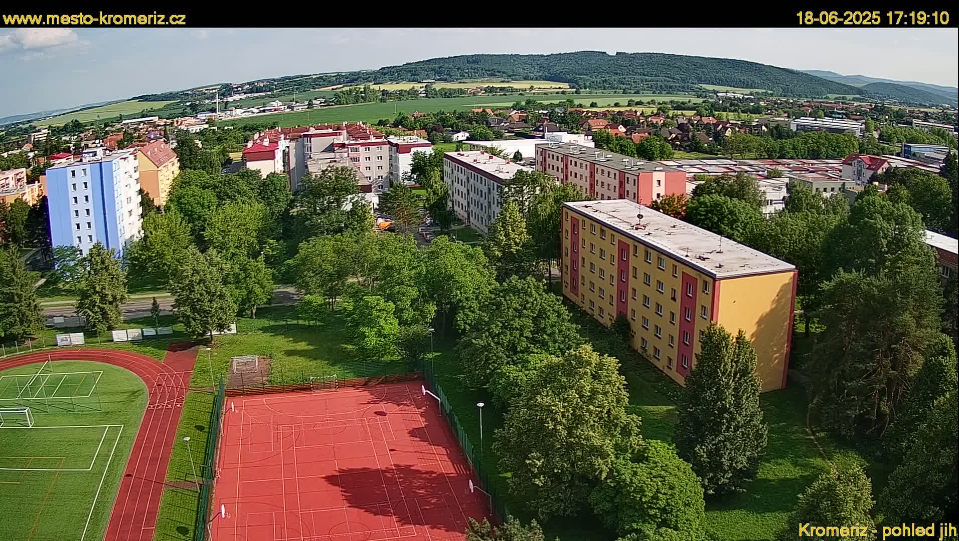 An aerial view shows a residential area with multi-story apartment buildings, green spaces, and athletic fields under a mostly sunny sky.