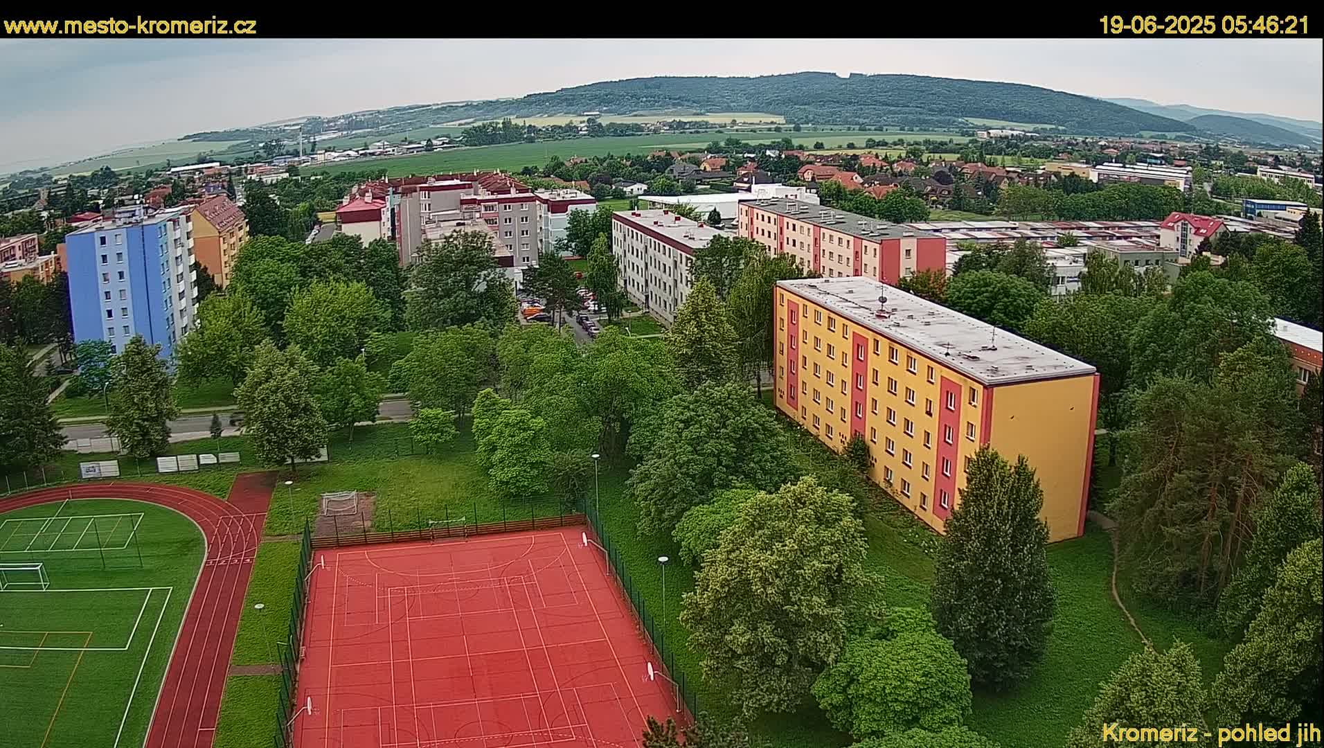 An aerial view shows a residential area with apartment buildings surrounded by lush green trees and a red sports court with a running track under an overcast sky.