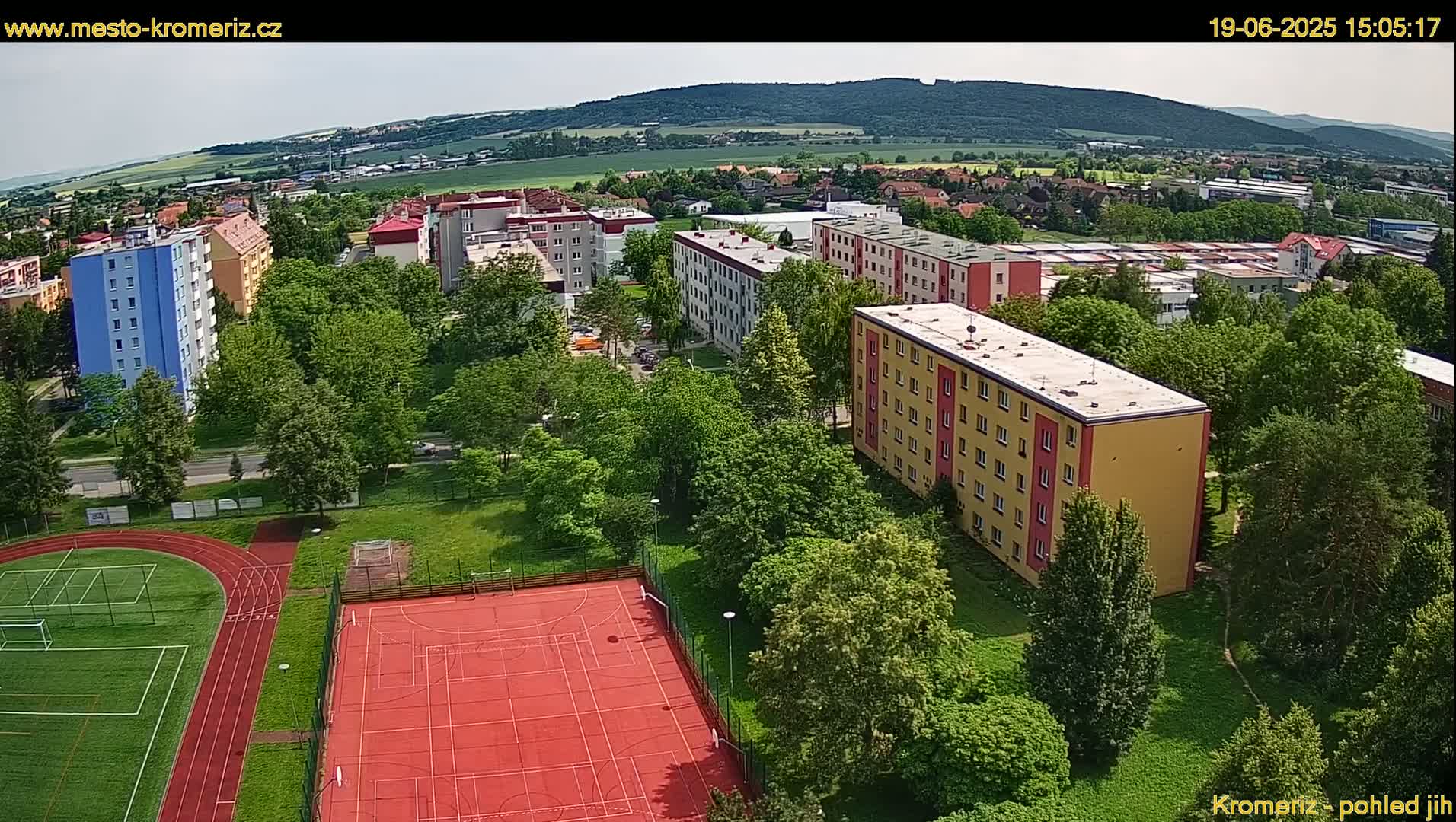 An aerial view shows apartment buildings surrounded by lush green trees and a red athletic field, all under a mostly clear sky.