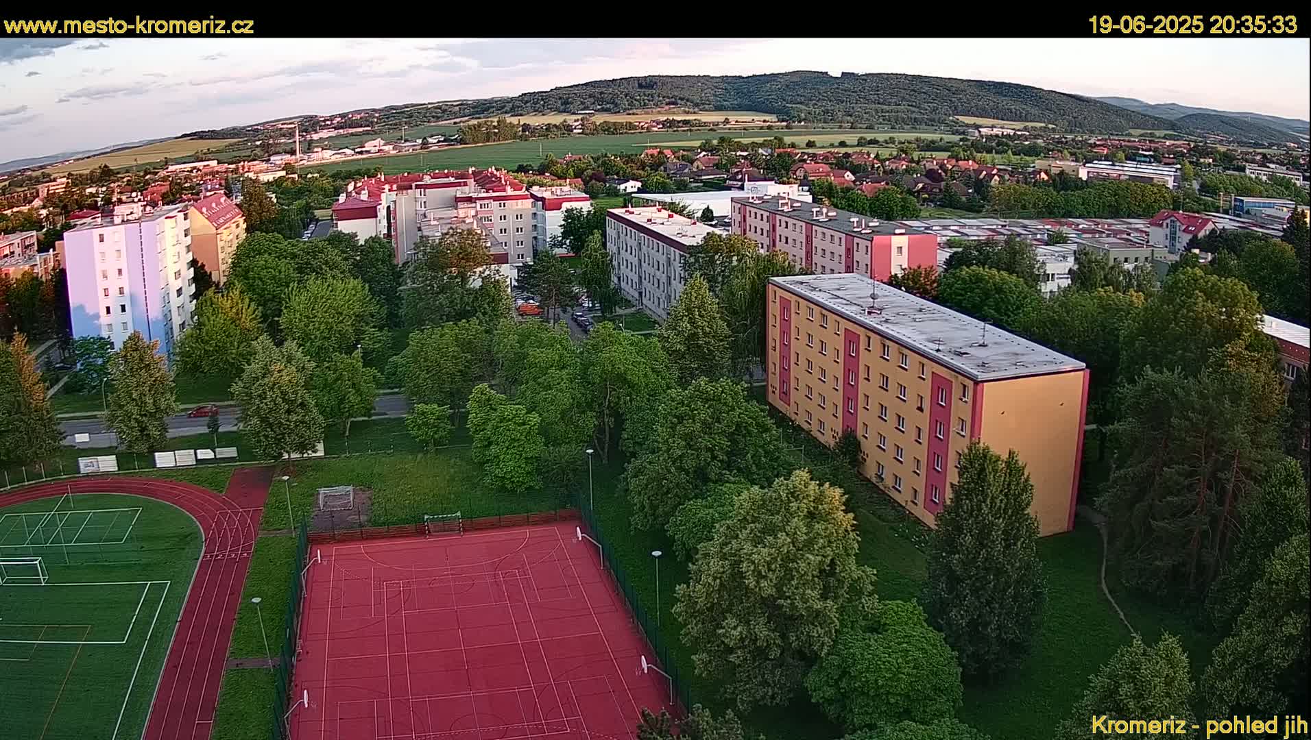 An aerial view of apartment buildings surrounded by lush green trees and a red sports court, under a partly cloudy evening sky.