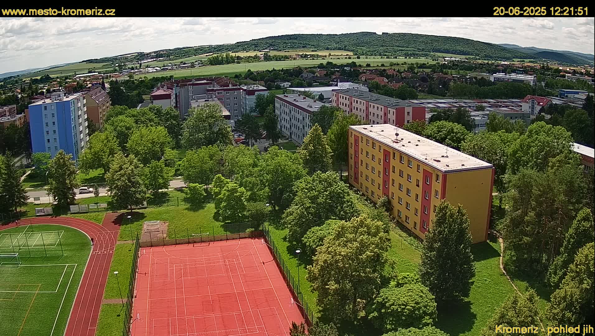 An aerial view shows a residential area with multi-story buildings, green spaces including a park and a sports field with a running track and tennis courts, under a partly cloudy sky.