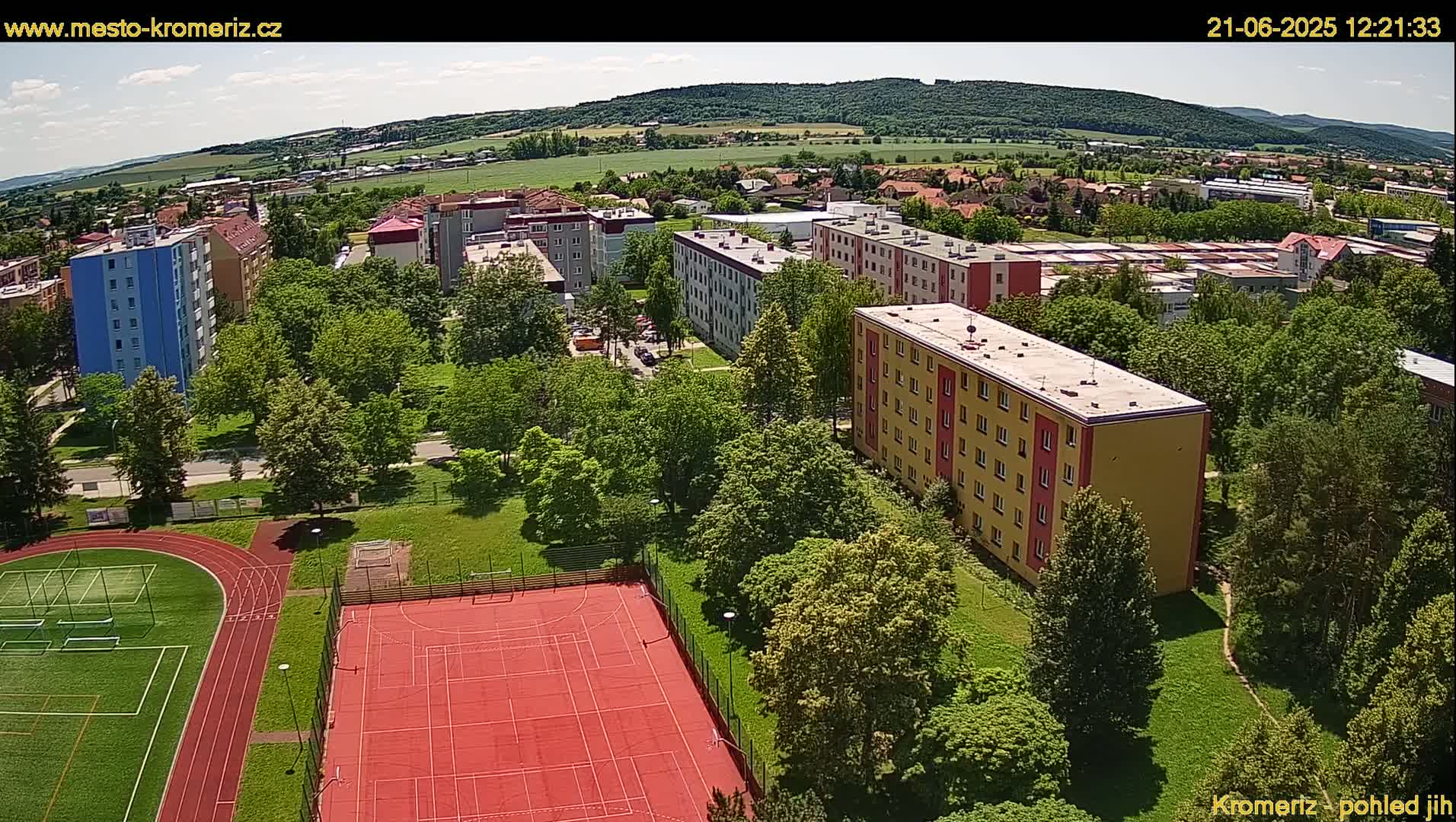 An aerial view of apartment buildings and a sports field surrounded by lush green trees on a sunny day.