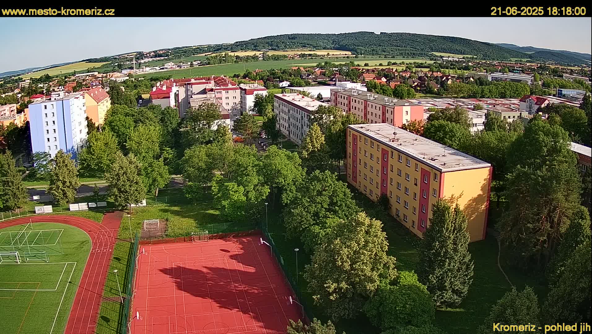 An aerial view shows a residential area with multi-story buildings and a sports field, under sunny skies.