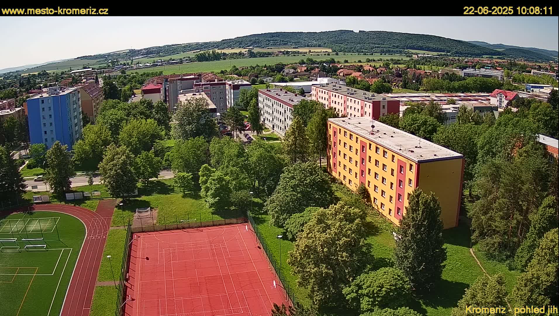An aerial view of apartment buildings and a sports field on a sunny day, with green trees and hills in the background.