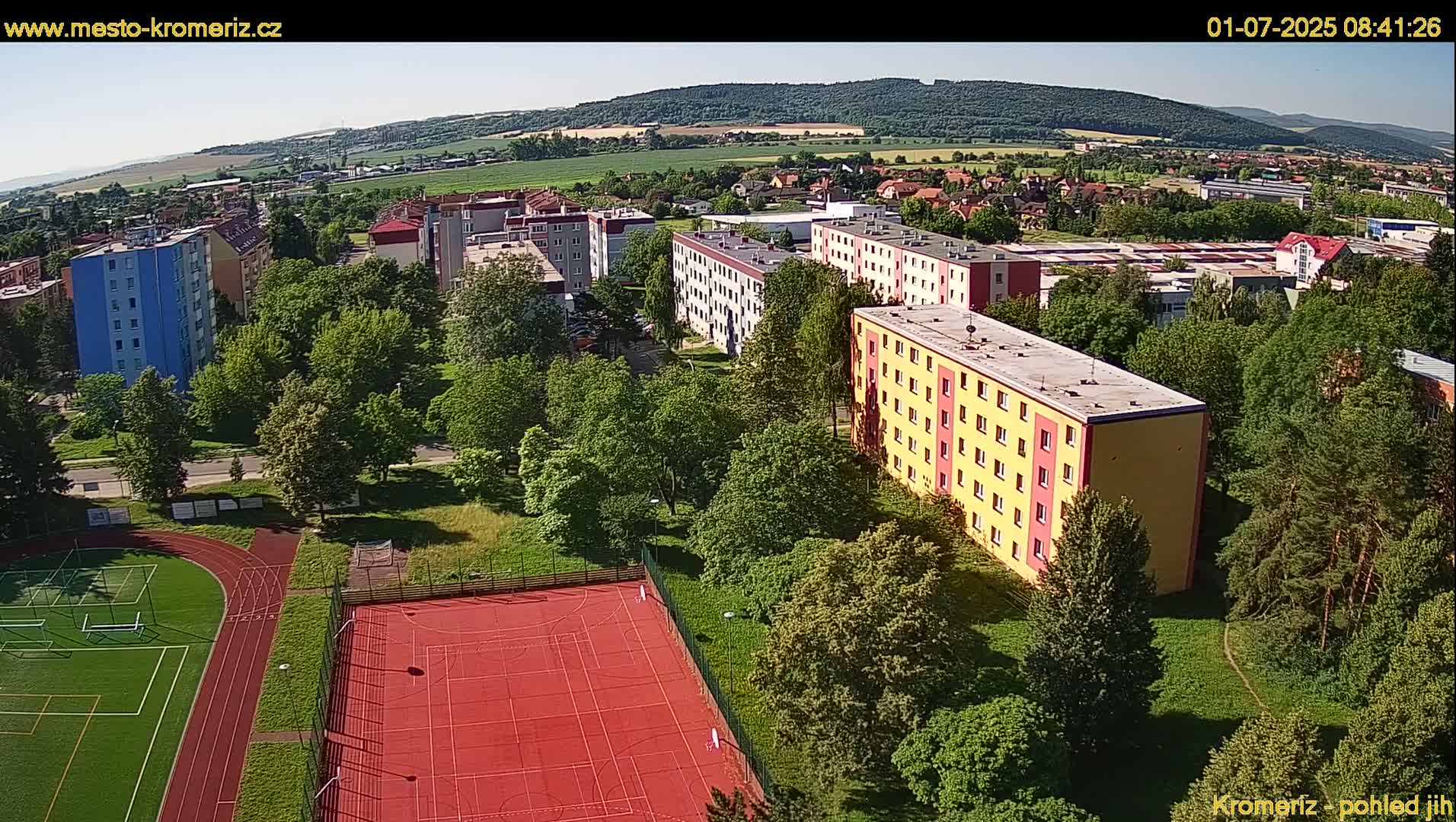An aerial view shows apartment buildings, green spaces with trees, and a red athletic field, all under a sunny sky.